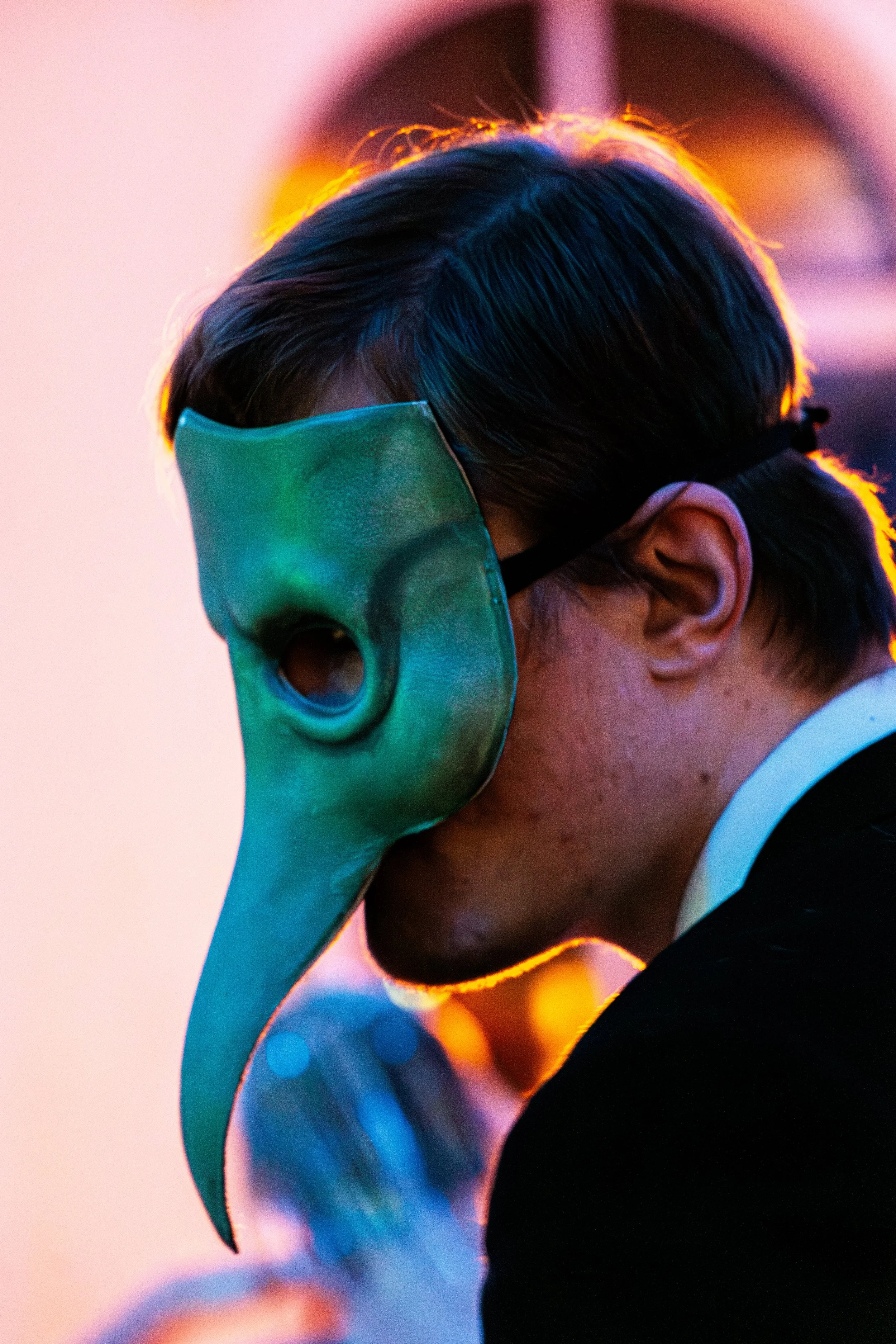 A person wearing a teal-colored mask with eye and beak details, resembling a bird mask, and a formal black suit.