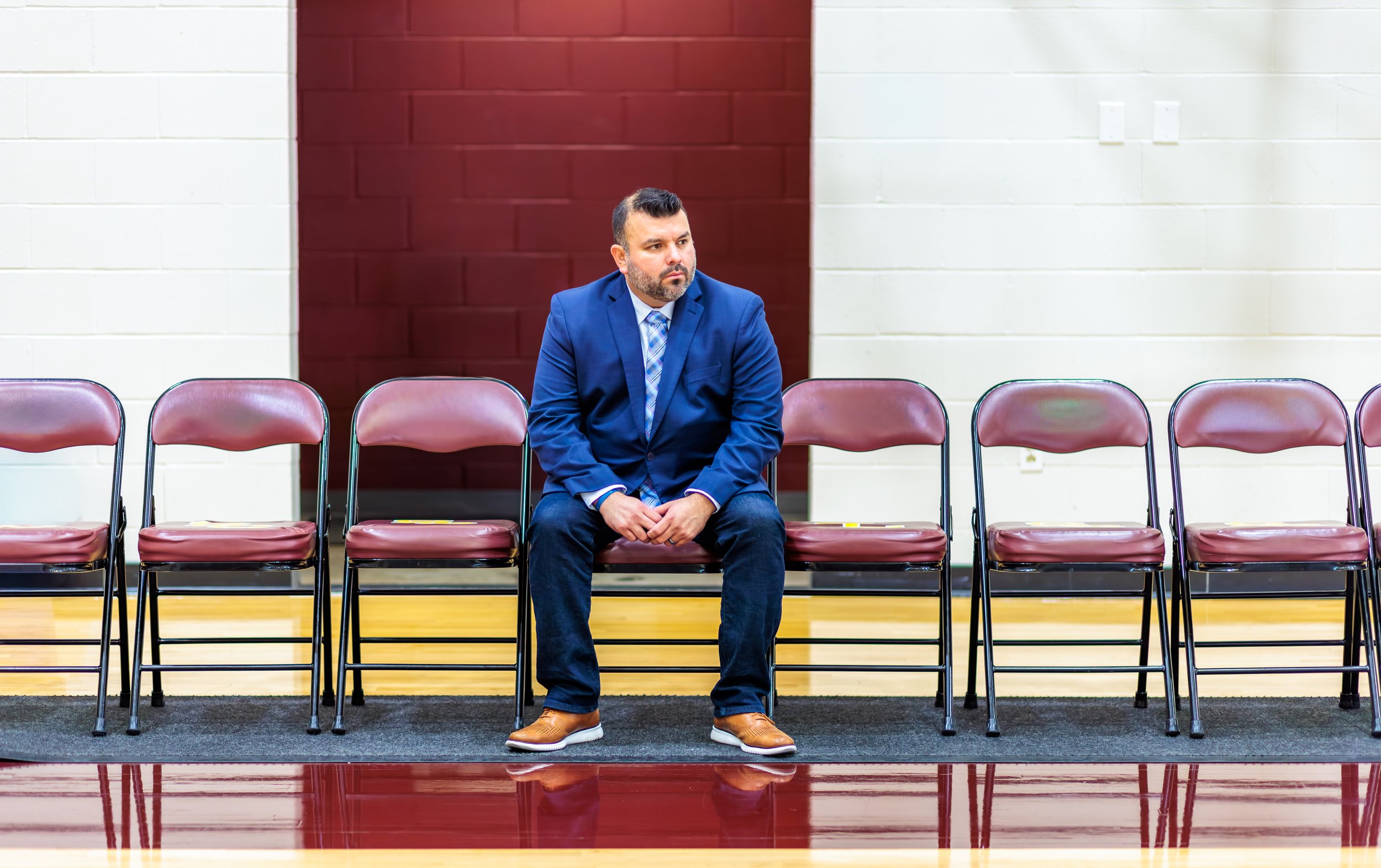 A man in a blue suit and tan shoes sitting alone on a row of maroon chairs in a gymnasium or auditorium, with a serious expression.