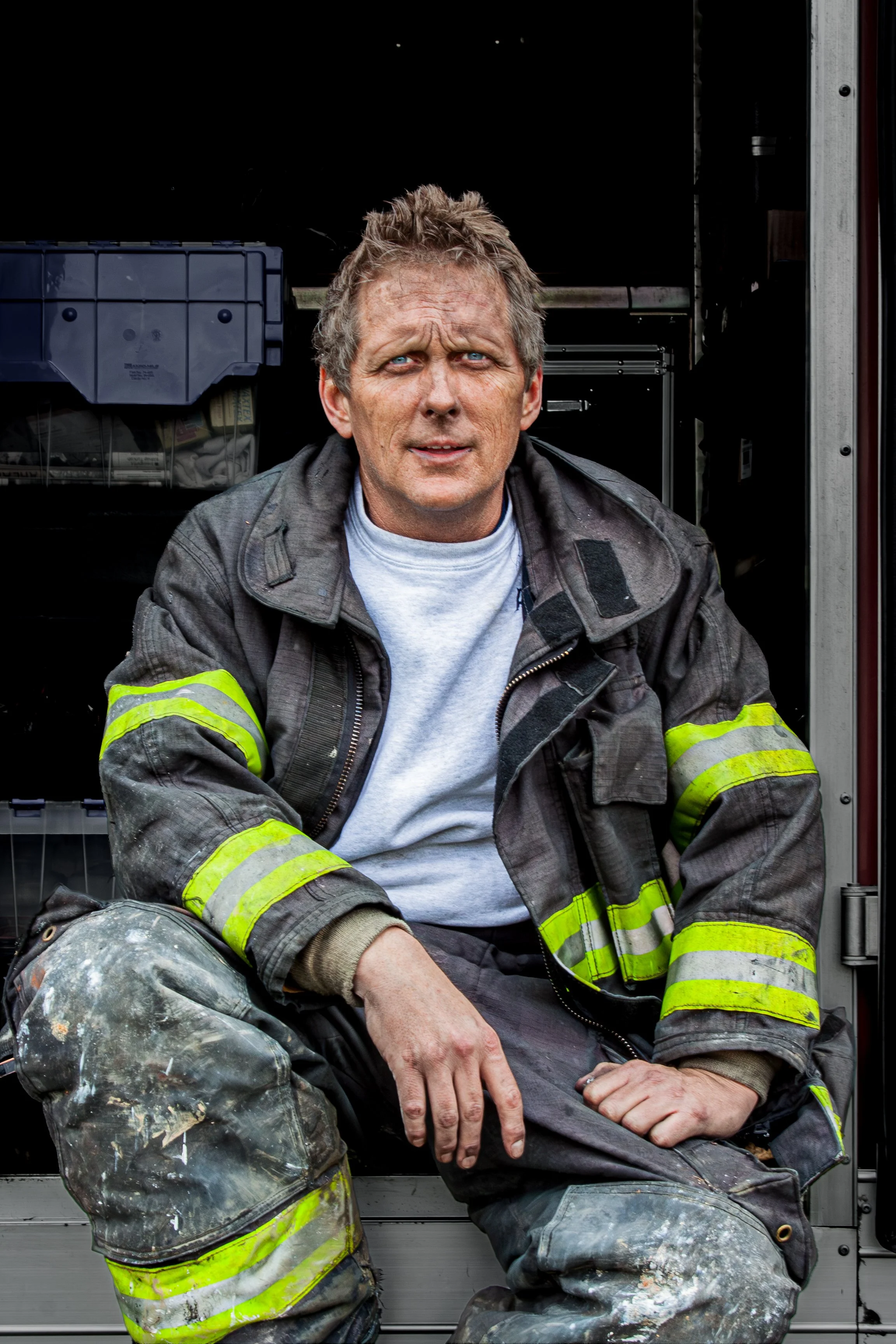 A middle-aged man in firefighter gear, including a black jacket with reflective stripes, sitting with his hand on his knee, looking into the camera with a serious expression. He has short, tousled hair and blue eyes. The background shows part of a ve