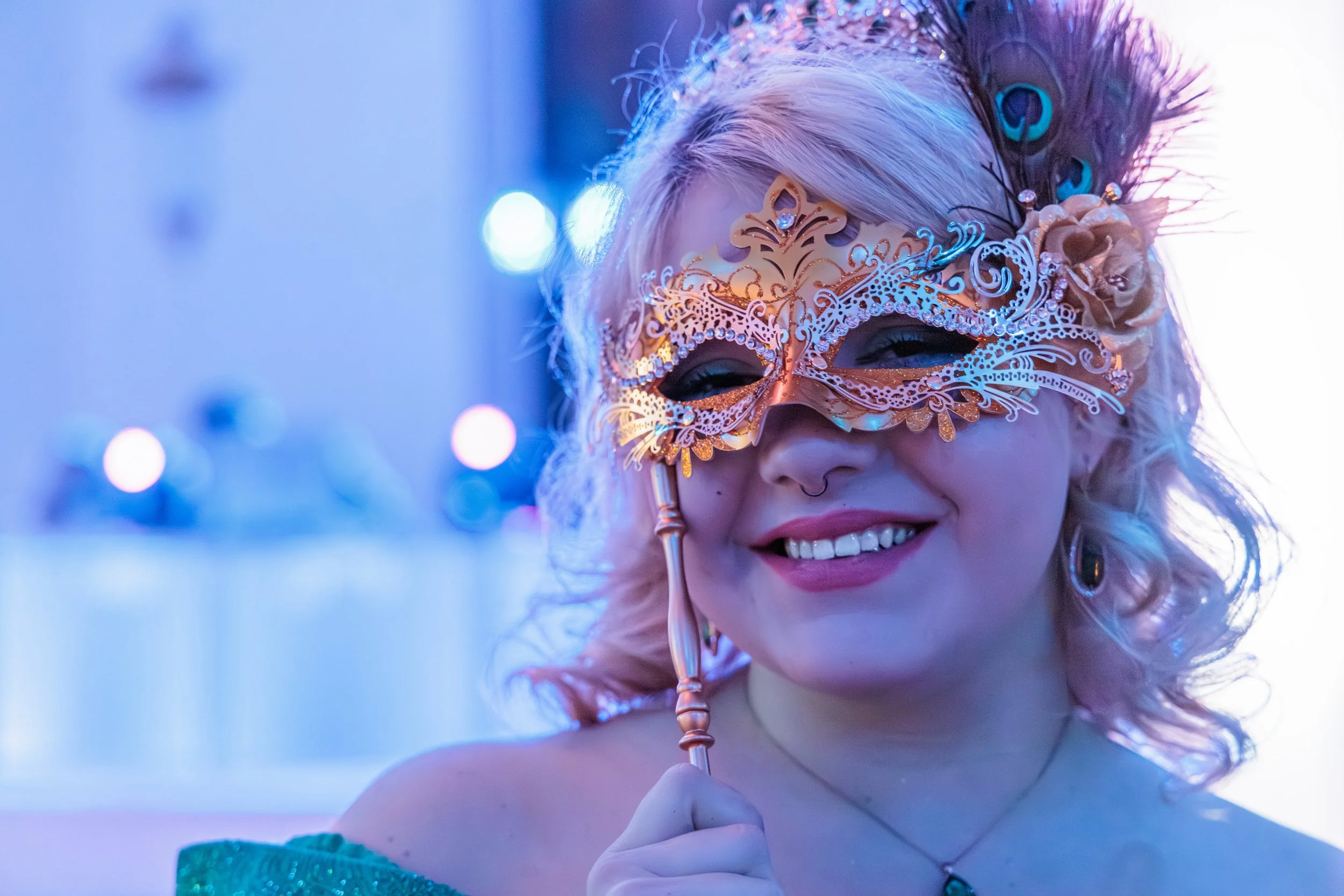 A young woman with pink curly hair holding a gold ornate masquerade mask with peacock feather details near her face, smiling in a festive setting with purple and blue lighting.