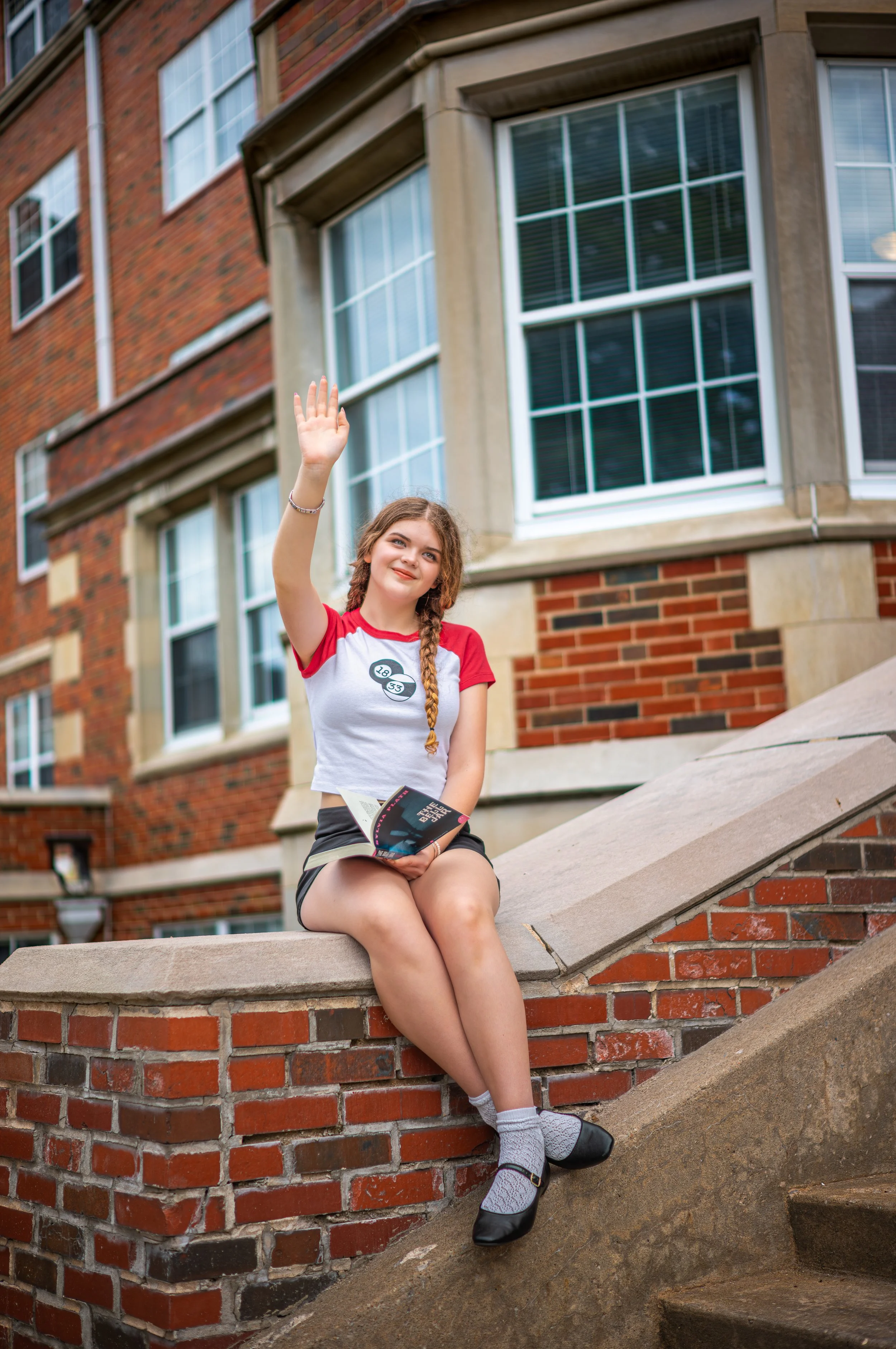 A young girl with braided hair, wearing a red and white t-shirt, shorts, and black shoes, sitting on stone steps in front of a red brick apartment building with large windows, waving.