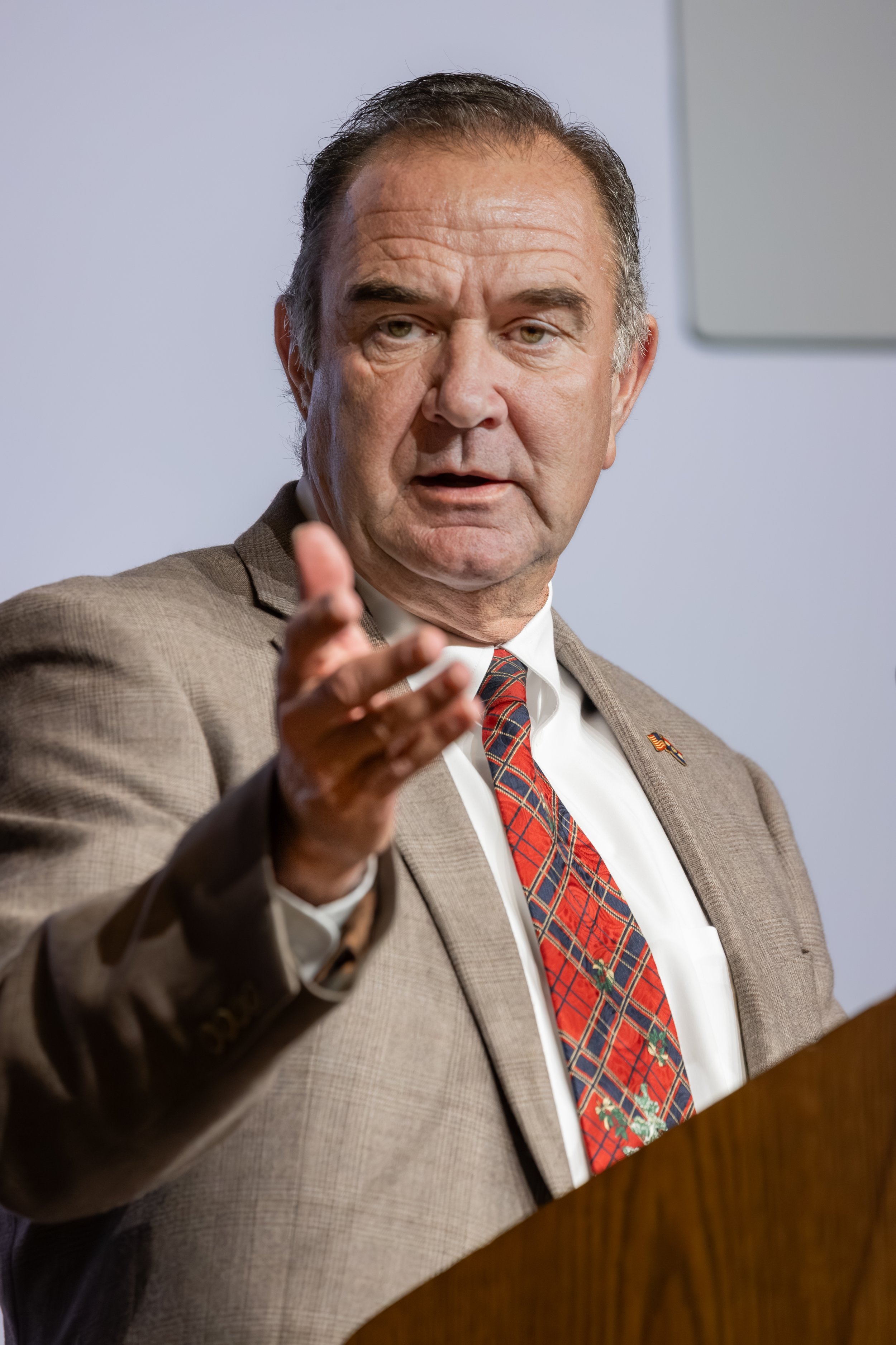 A middle-aged man with short dark hair and light skin, wearing a beige plaid suit, white shirt, and Christmas-themed tie, speaking at a podium with a serious expression.