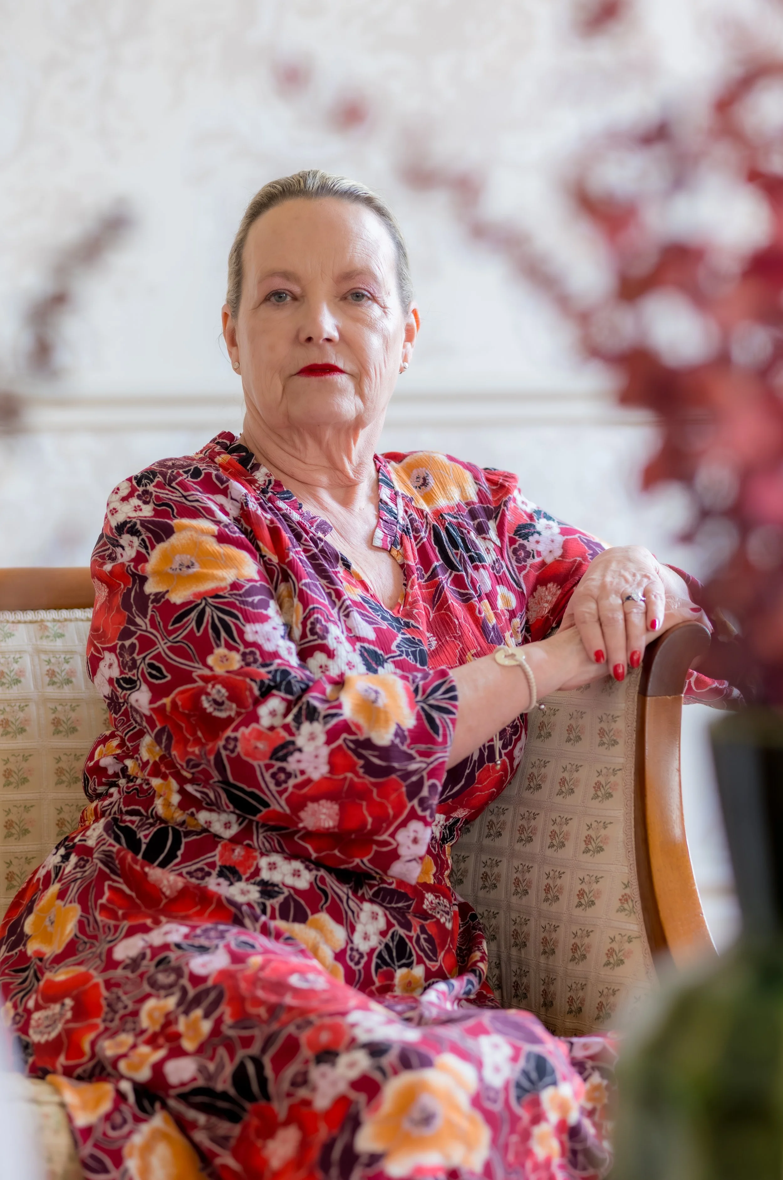 An elderly woman with gray hair, wearing red lipstick, and a colorful floral dress, sitting on a chair indoors, with a blurred floral background.