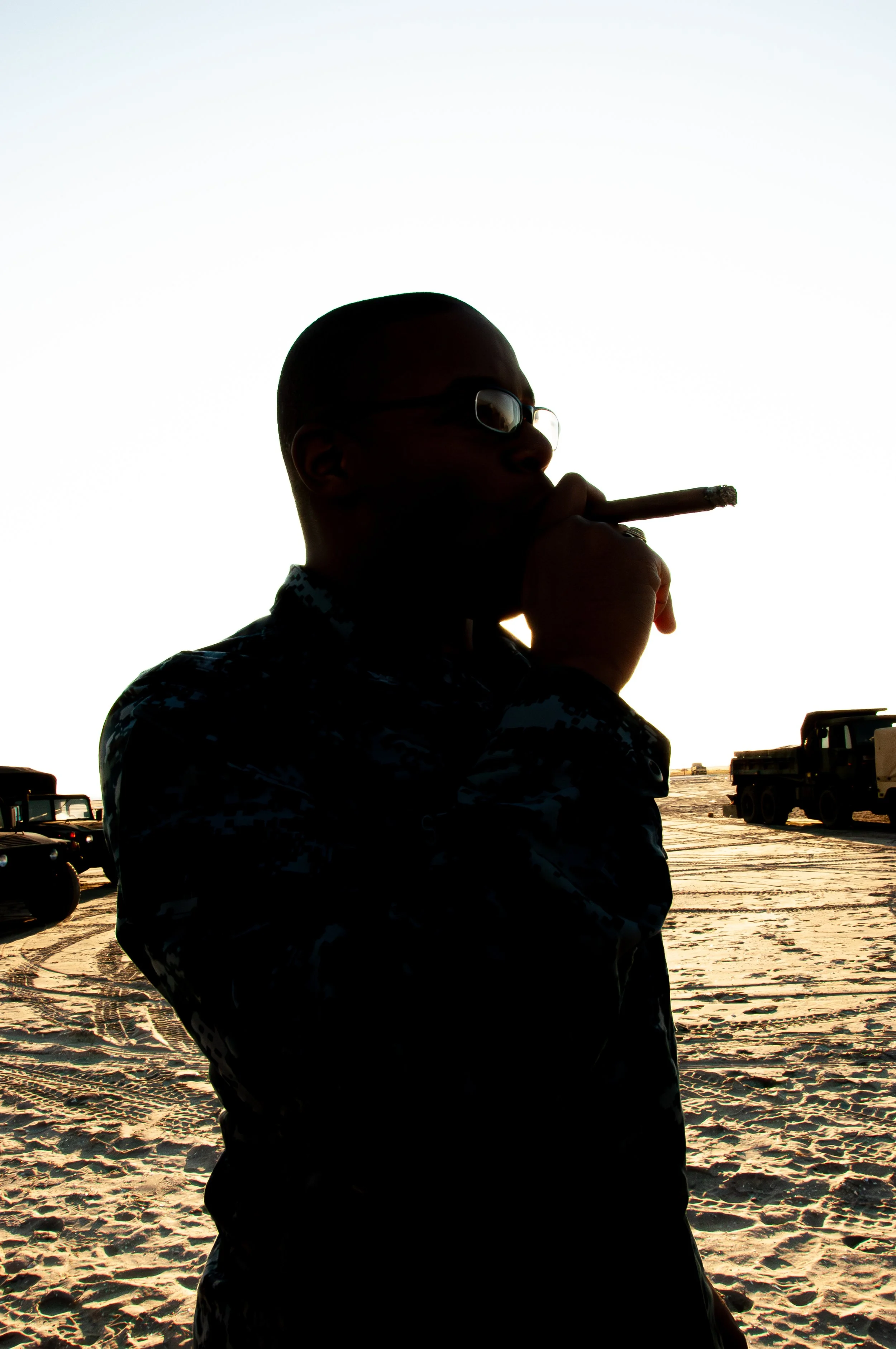 Silhouette of a man wearing glasses, smoking a cigar, standing on a beach with a sunset or sunrise in the background, and military trucks parked nearby.