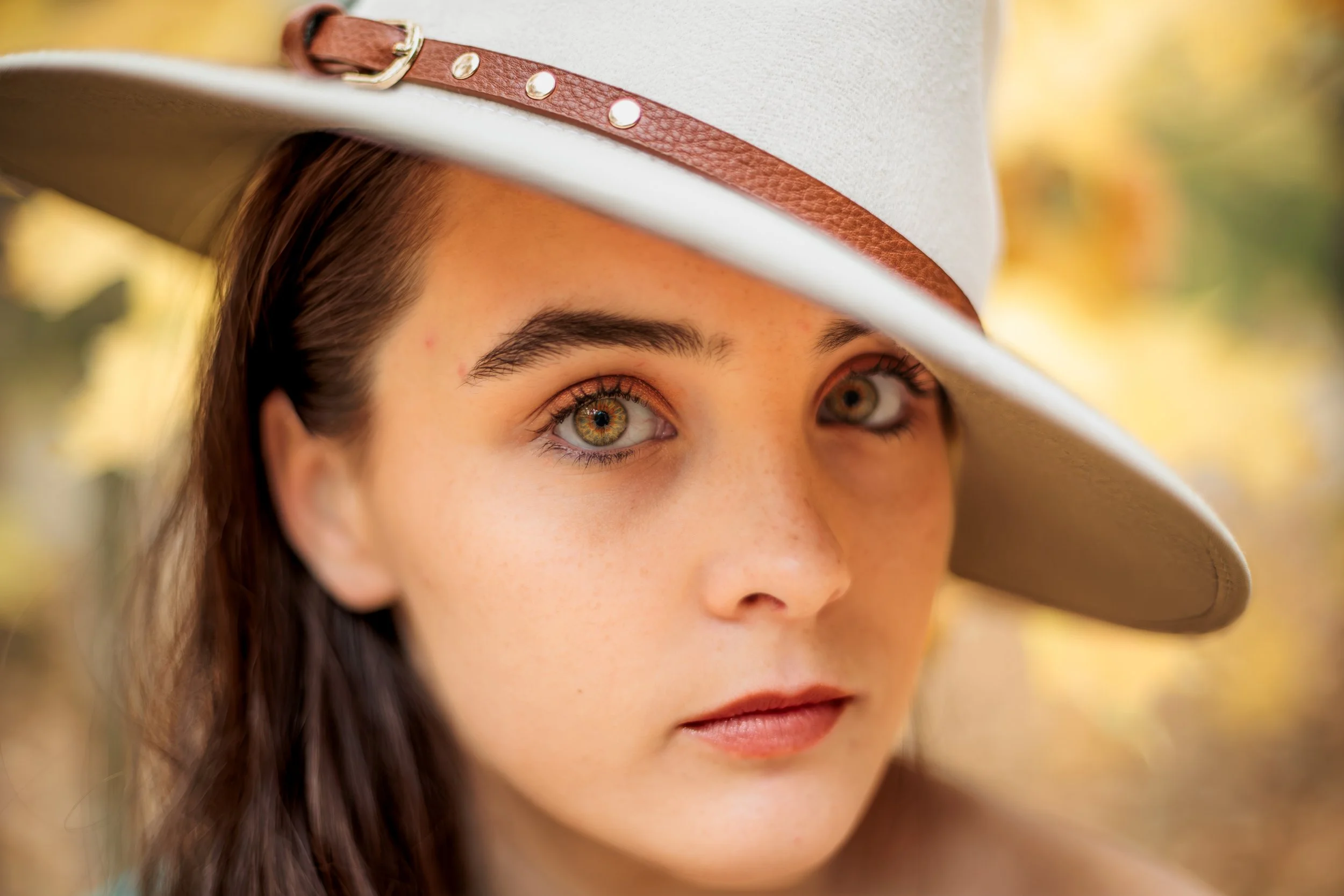 Close-up of a woman with brown hair wearing a light-colored cowboy hat with brown leather band, outdoors with blurred yellow and green background.
