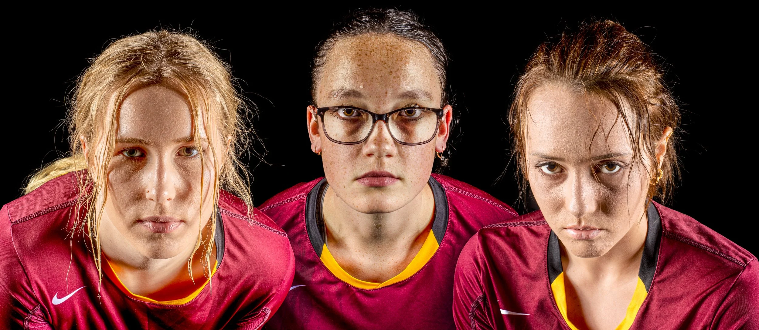 Three female athletes wearing maroon sports jerseys looking directly at the camera against a black background.