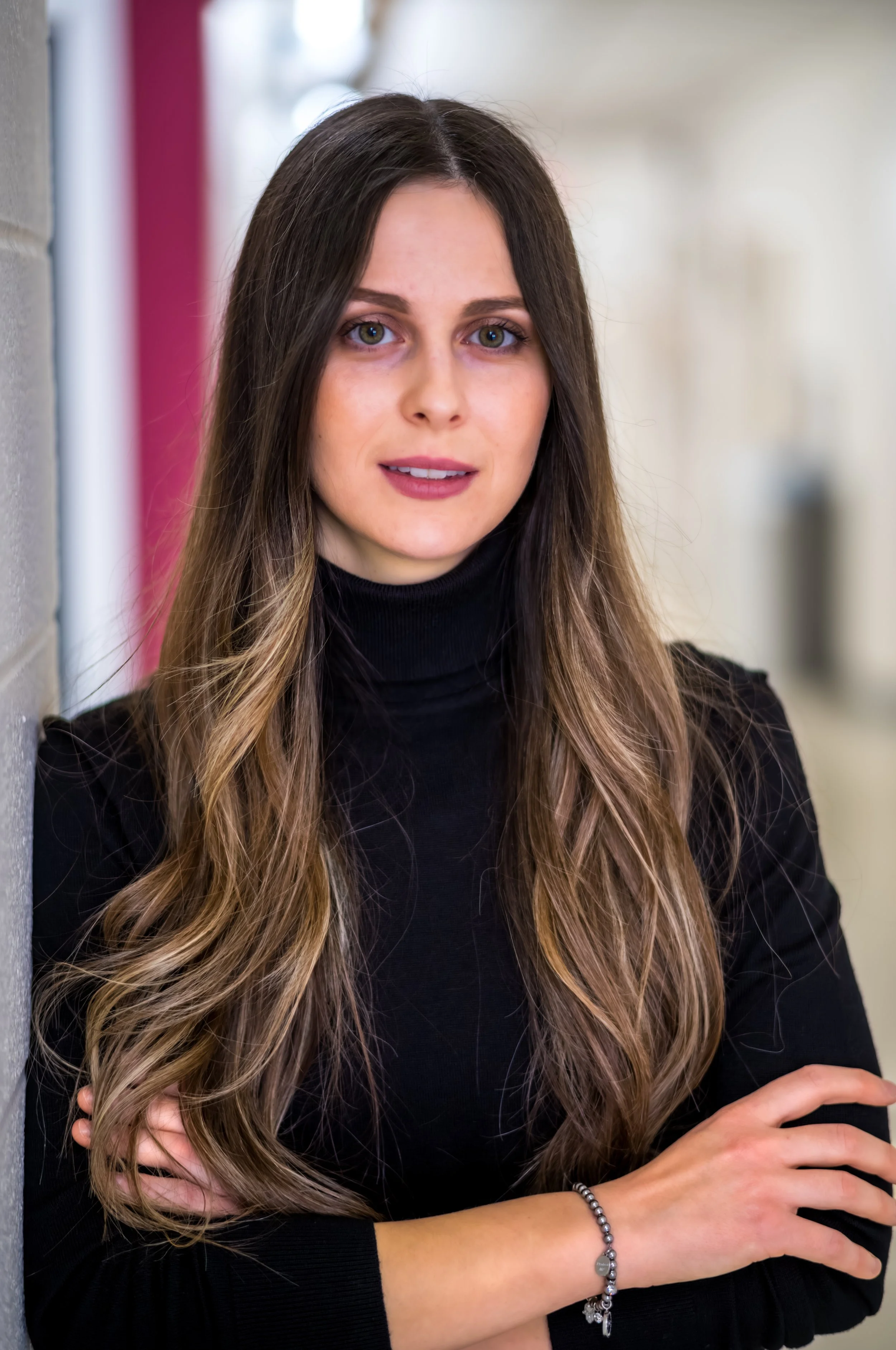 A woman with long, wavy brown hair and fair skin standing against a blurred background, wearing a black turtleneck and a bracelet on her wrist.
