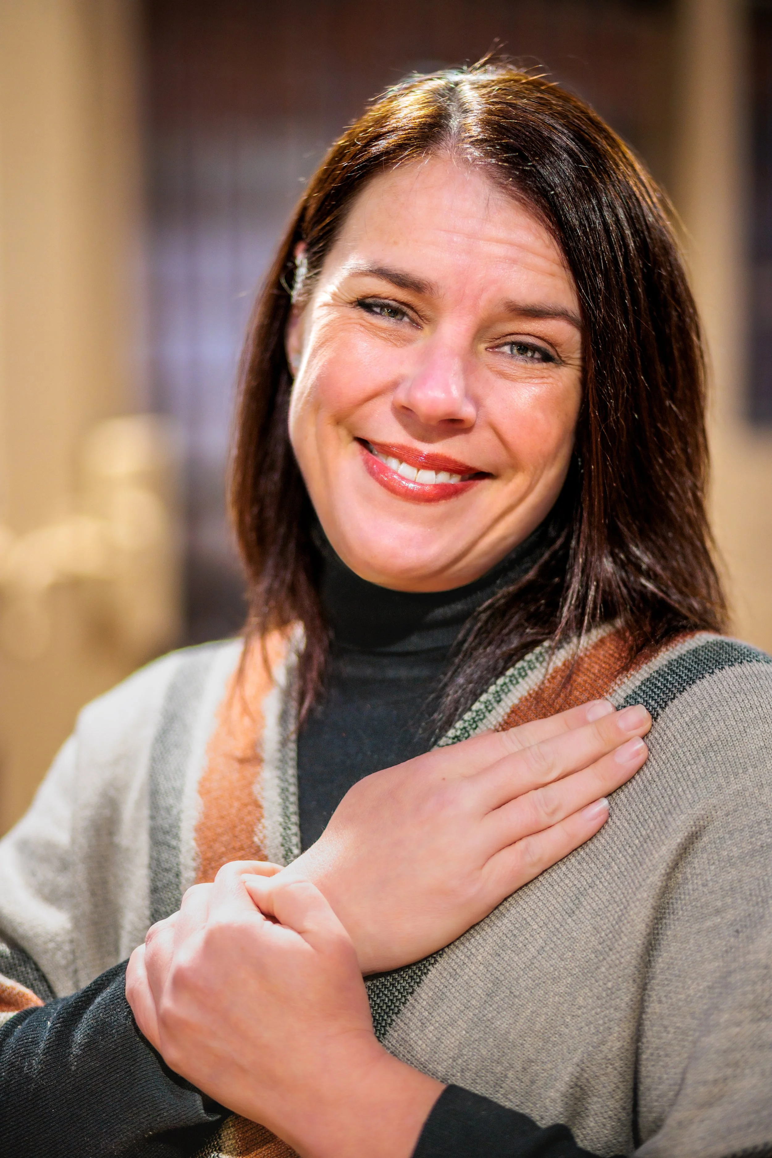 A woman with dark brown hair, wearing a black turtleneck and a beige cardigan with a multicolored stripe, smiling and touching her shoulder with one hand.