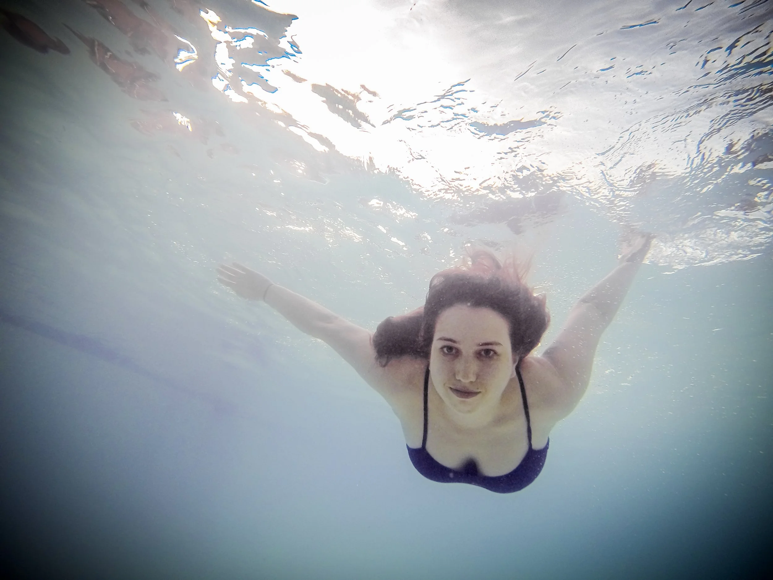 A young woman swimming underwater in a swimming pool, looking directly at the camera with a neutral expression, arms extended outward.