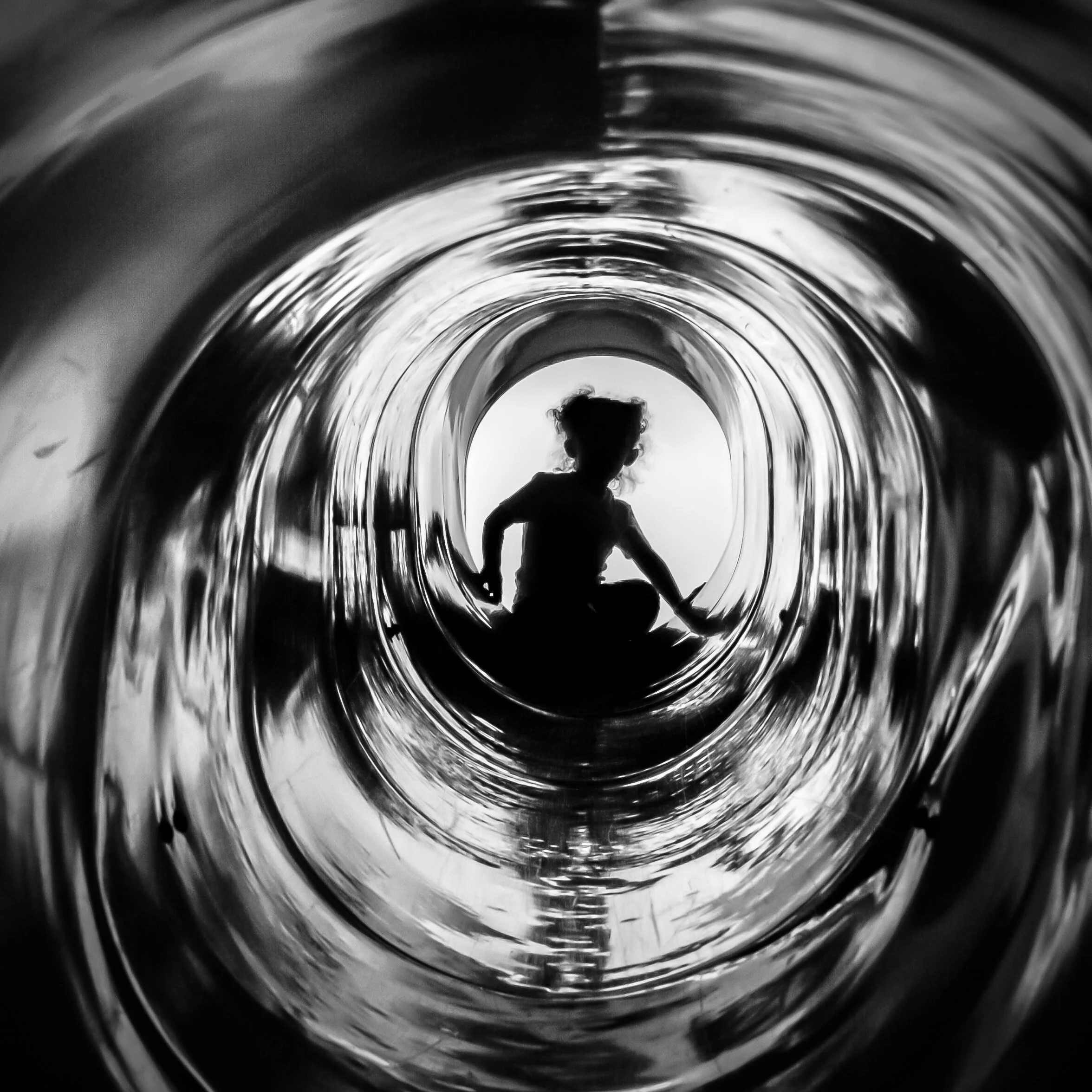 Black and white photo showing the inside of a bottle or tube, with a person standing at the entrance. The person appears to be a child with curly hair, wearing a shirt, seen from behind.
