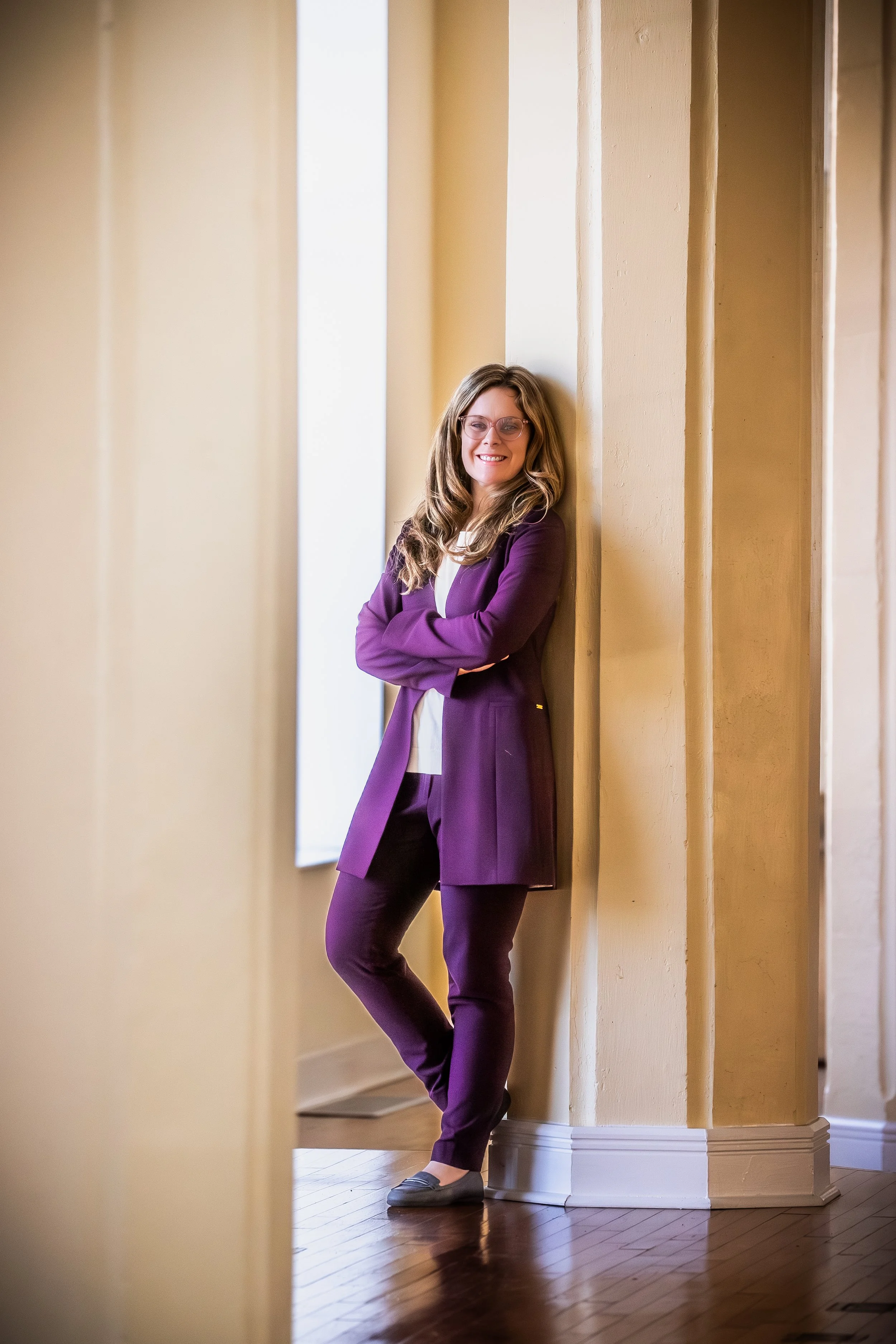 A smiling woman with glasses, wearing a purple suit, standing with arms crossed against a yellow wall in a bright indoor space.