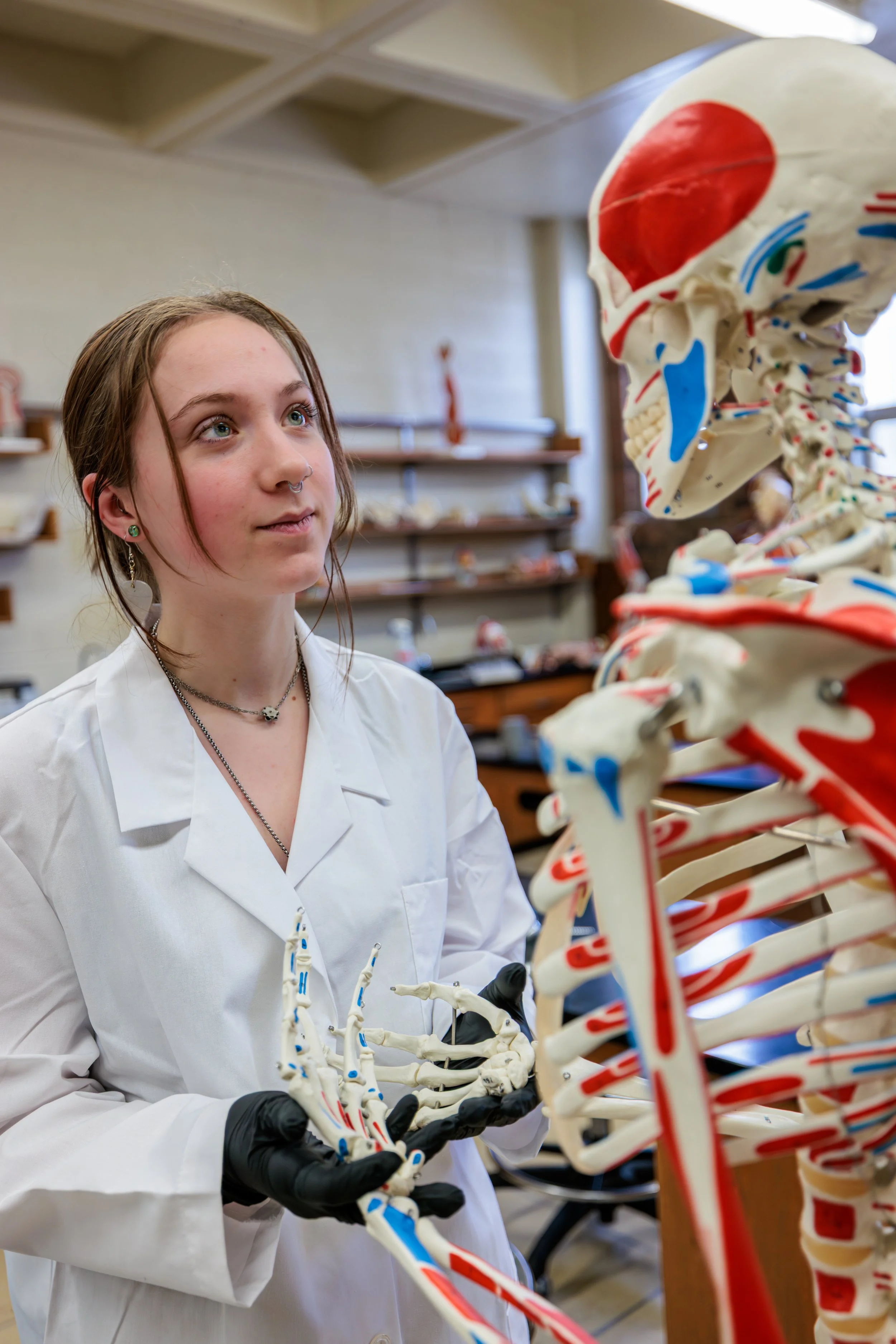 A young woman with brown hair, wearing a white lab coat, black gloves, and a septum piercing, looks at a human skeleton model in a classroom or laboratory setting.