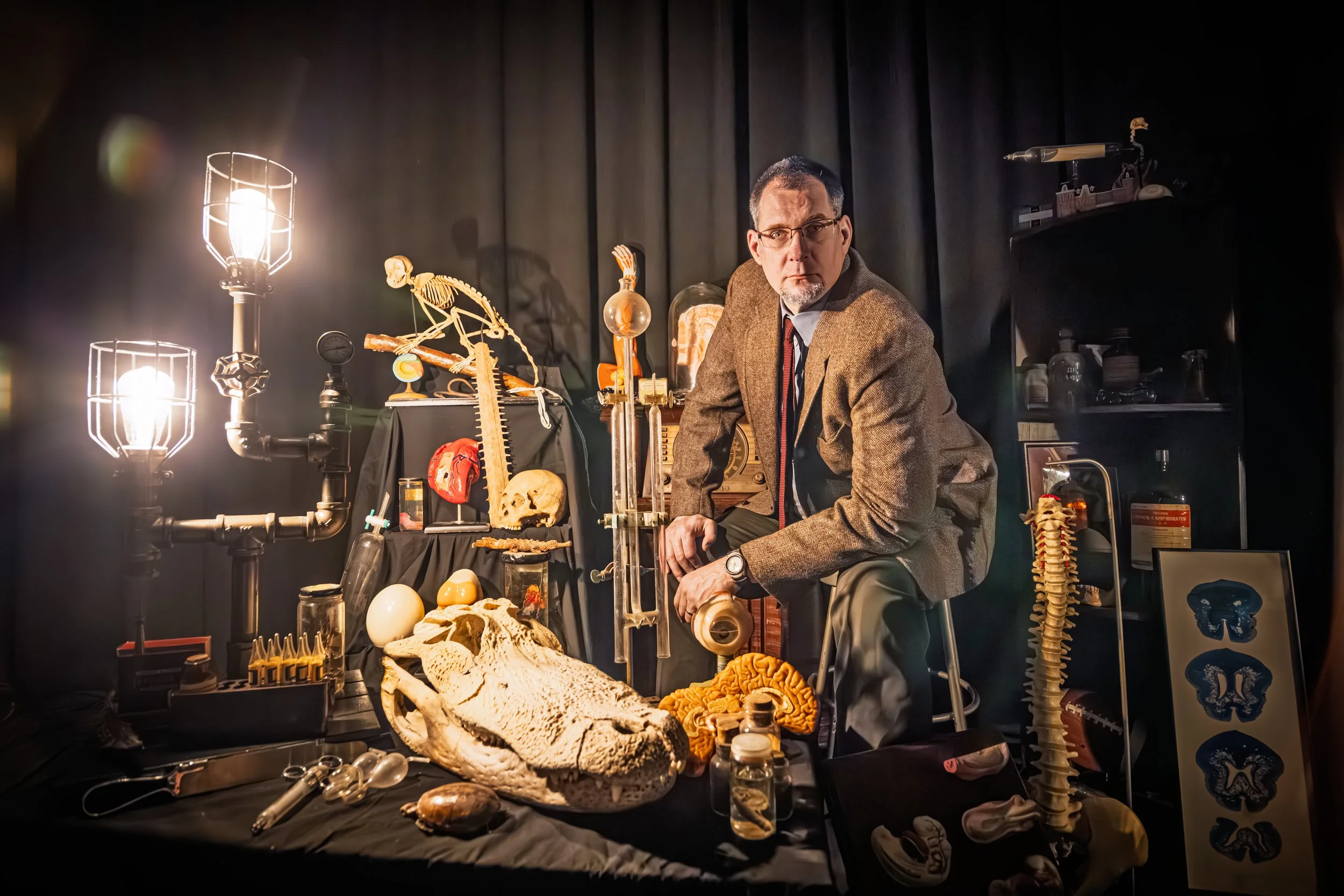 A scientist in a brown blazer and glasses sitting among various scientific models and equipment in a laboratory setting. There are anatomical skulls, a spine model, light fixtures, and scientific instruments on and around the black table and shelves.