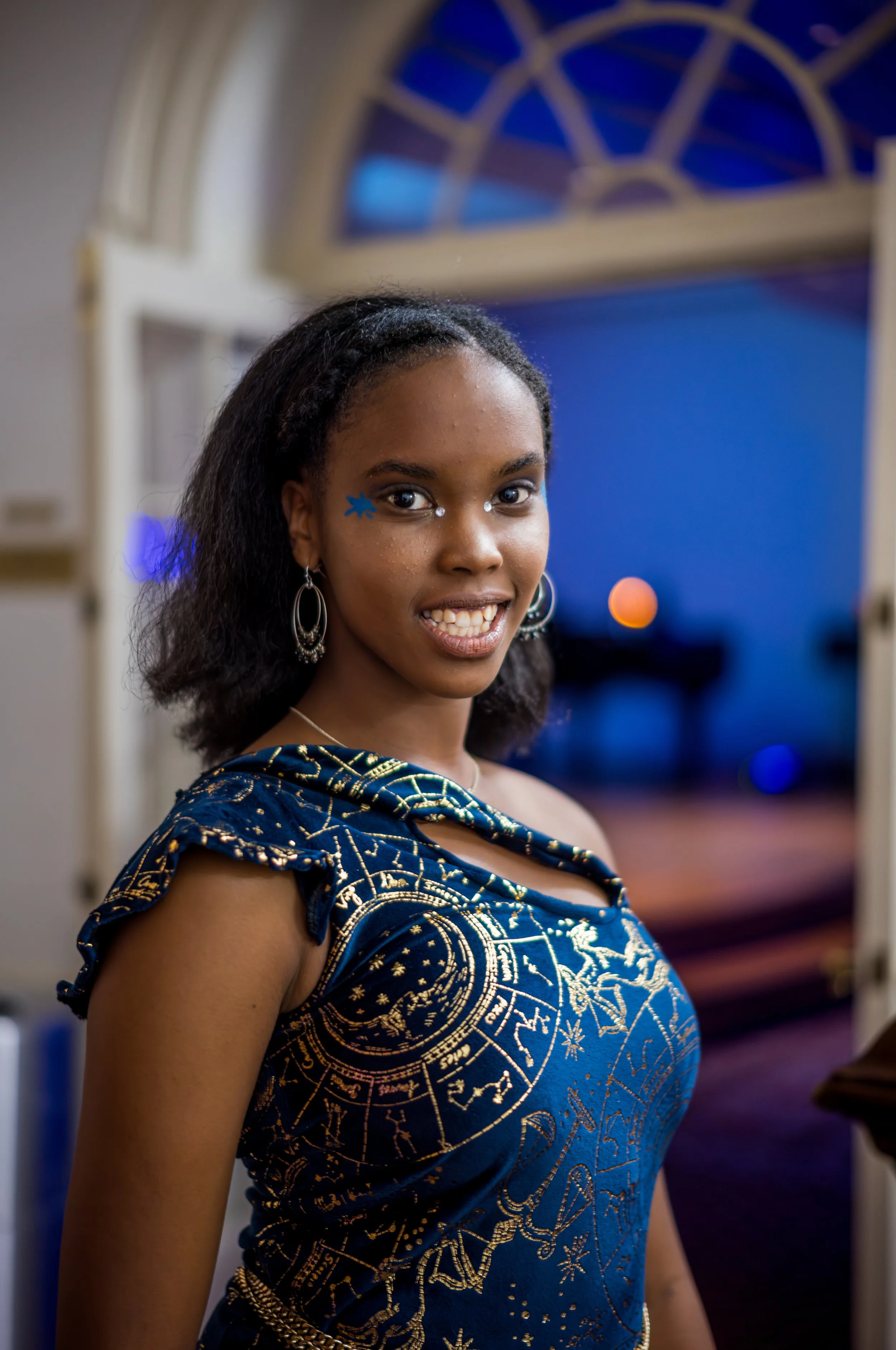 A young woman smiling at the camera, wearing a dark blue dress with a gold constellation and zodiac pattern, standing in an indoor setting with a large arched window behind her.