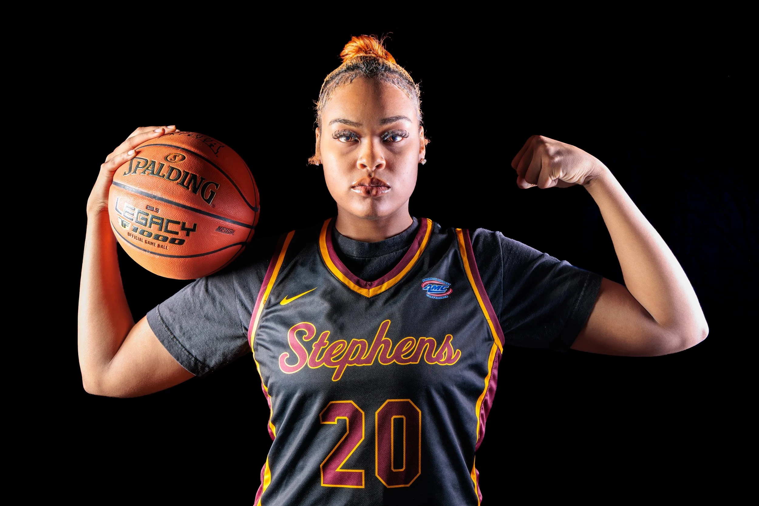 A young woman in a basketball jersey labeled 'Stephens' and number '20' is holding a basketball in her left hand and flexing her right arm, standing against a black background.