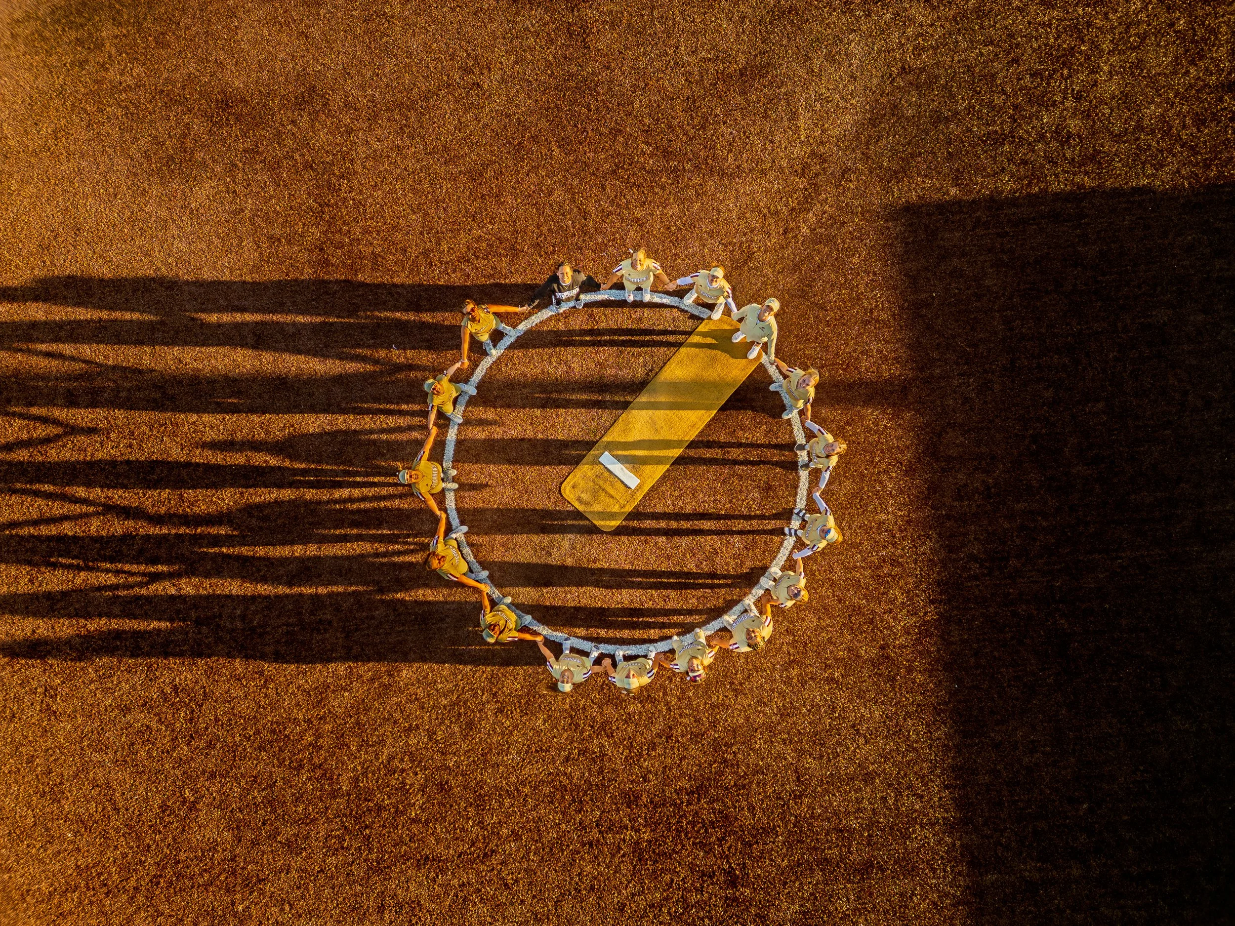 An aerial view of a group of football players sitting in a circle on a field with a yellow marker and long shadows.
