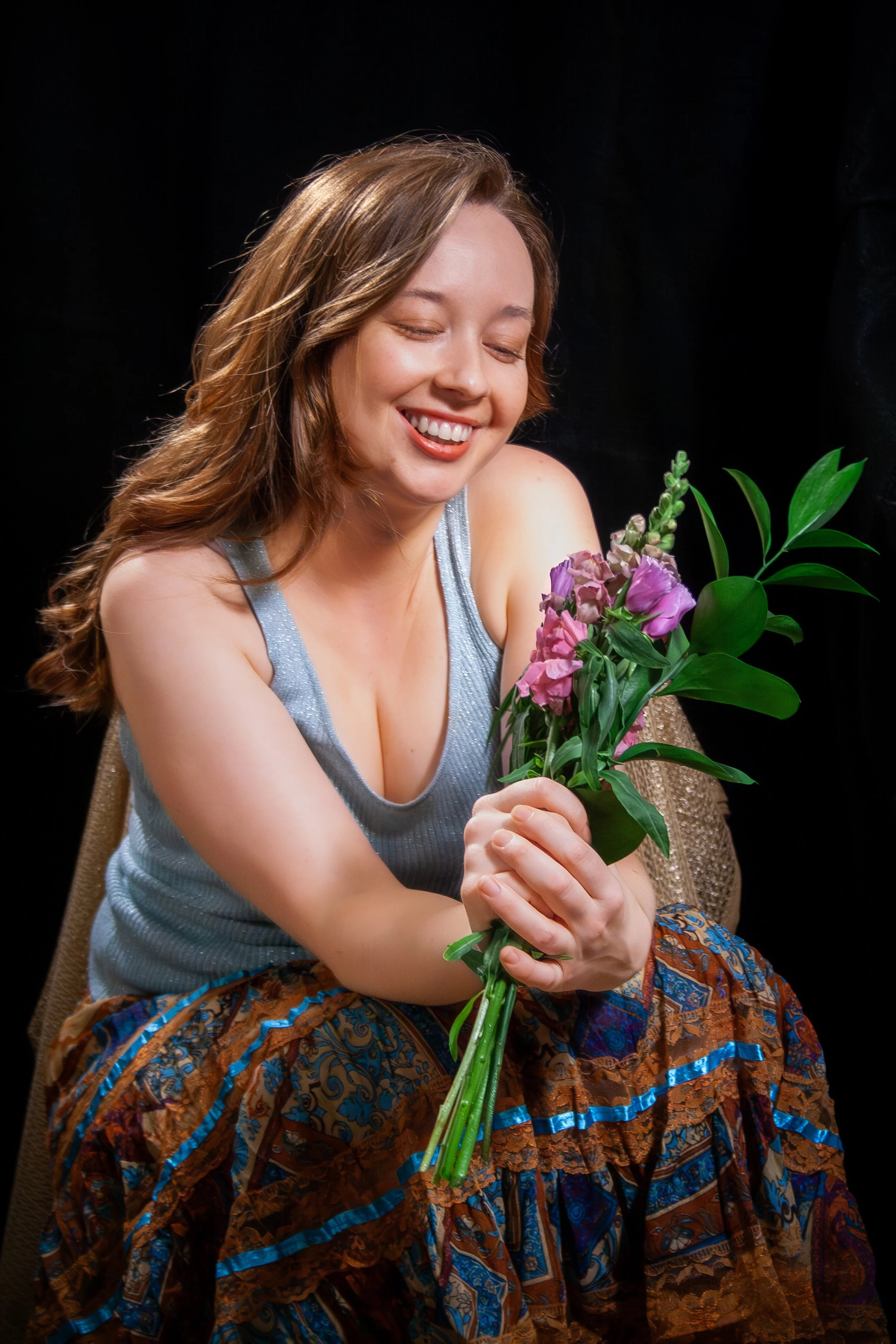 A woman with brown hair smiling and sitting on a chair, holding a bouquet of purple and pink flowers, against a dark background.