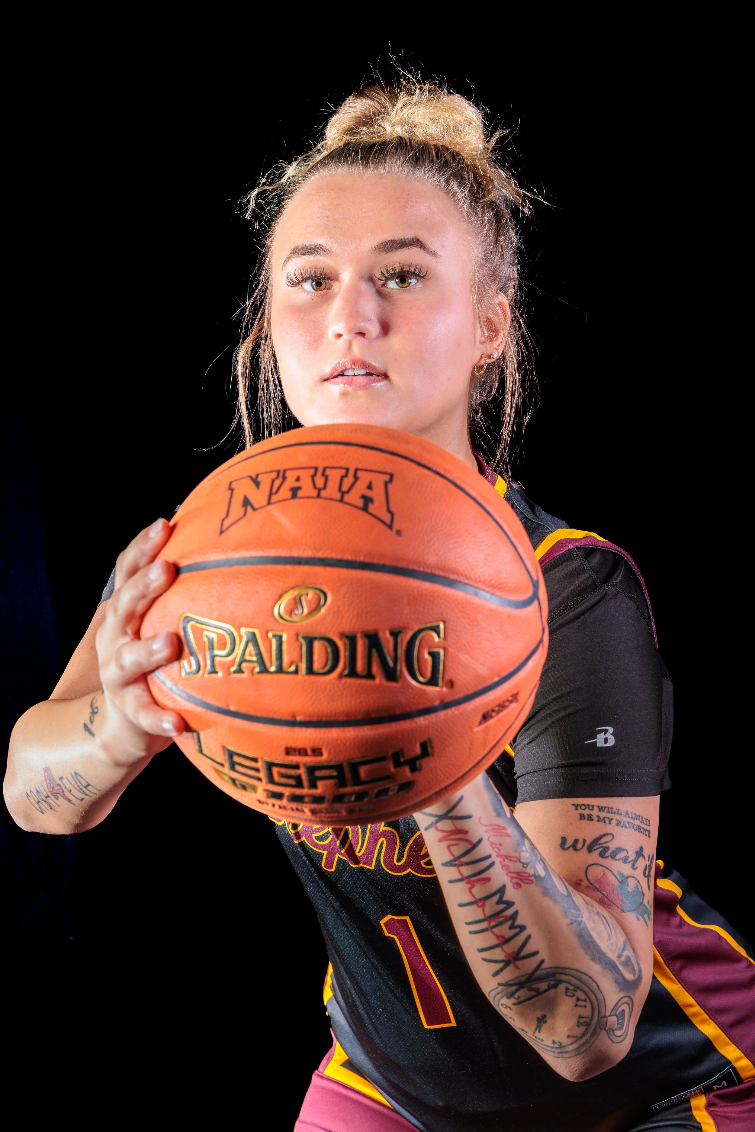 A female athlete with tattoos holding a basketball and preparing to shoot, wearing a black and maroon sports jersey against a black background.