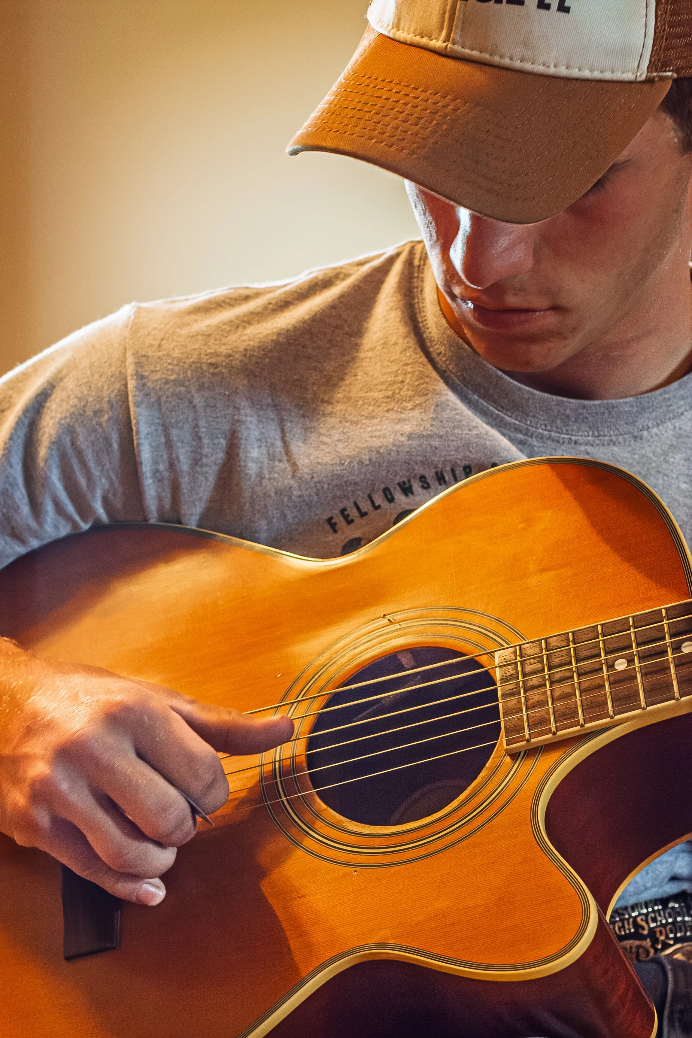 A person wearing a beige and brown baseball cap and a light gray T-shirt playing an acoustic guitar. The individual is focused on strumming the guitar with their right hand, and the image captures the upper body and part of the guitar.