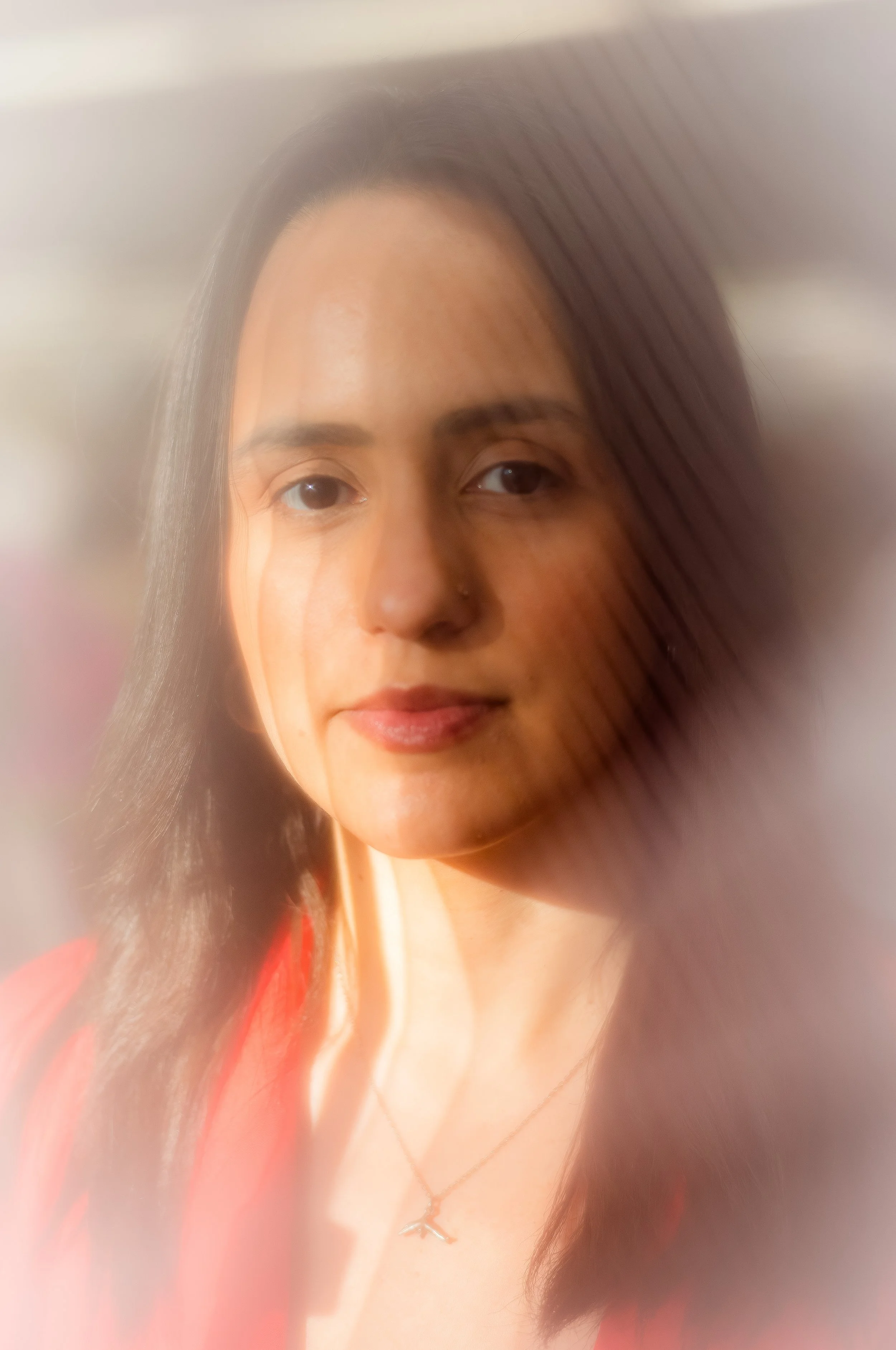 A close-up portrait of a woman with long dark hair, wearing a red top and a necklace with a small cross pendant. The image has a blurred and soft-focus effect around the edges.