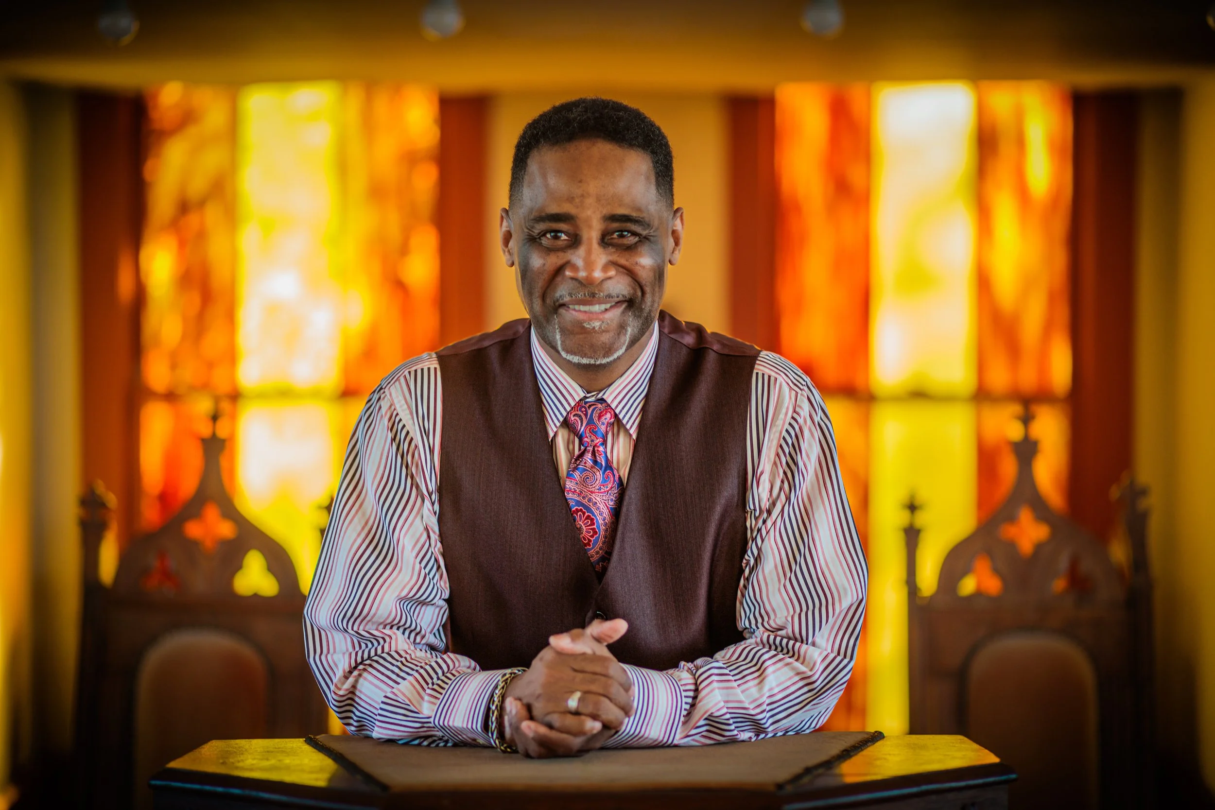 A man with dark skin, short black hair, and a grey beard, dressed in a striped shirt, a patterned tie, and a vest, sitting at a table with his hands clasped, smiling, with a colorful stained glass window background.