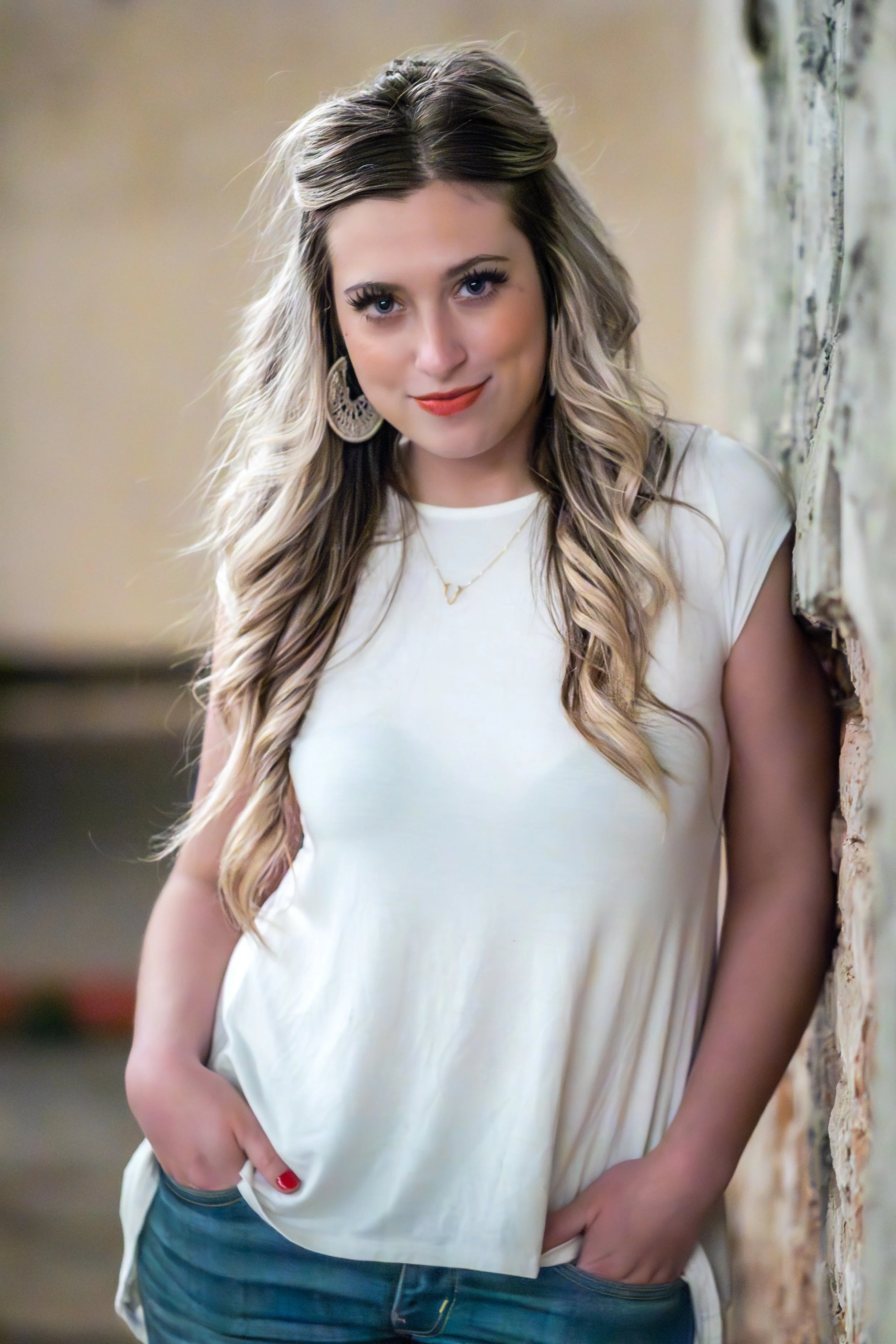 A woman with long, wavy blonde hair wearing a white top and large earrings, standing against a brick wall.