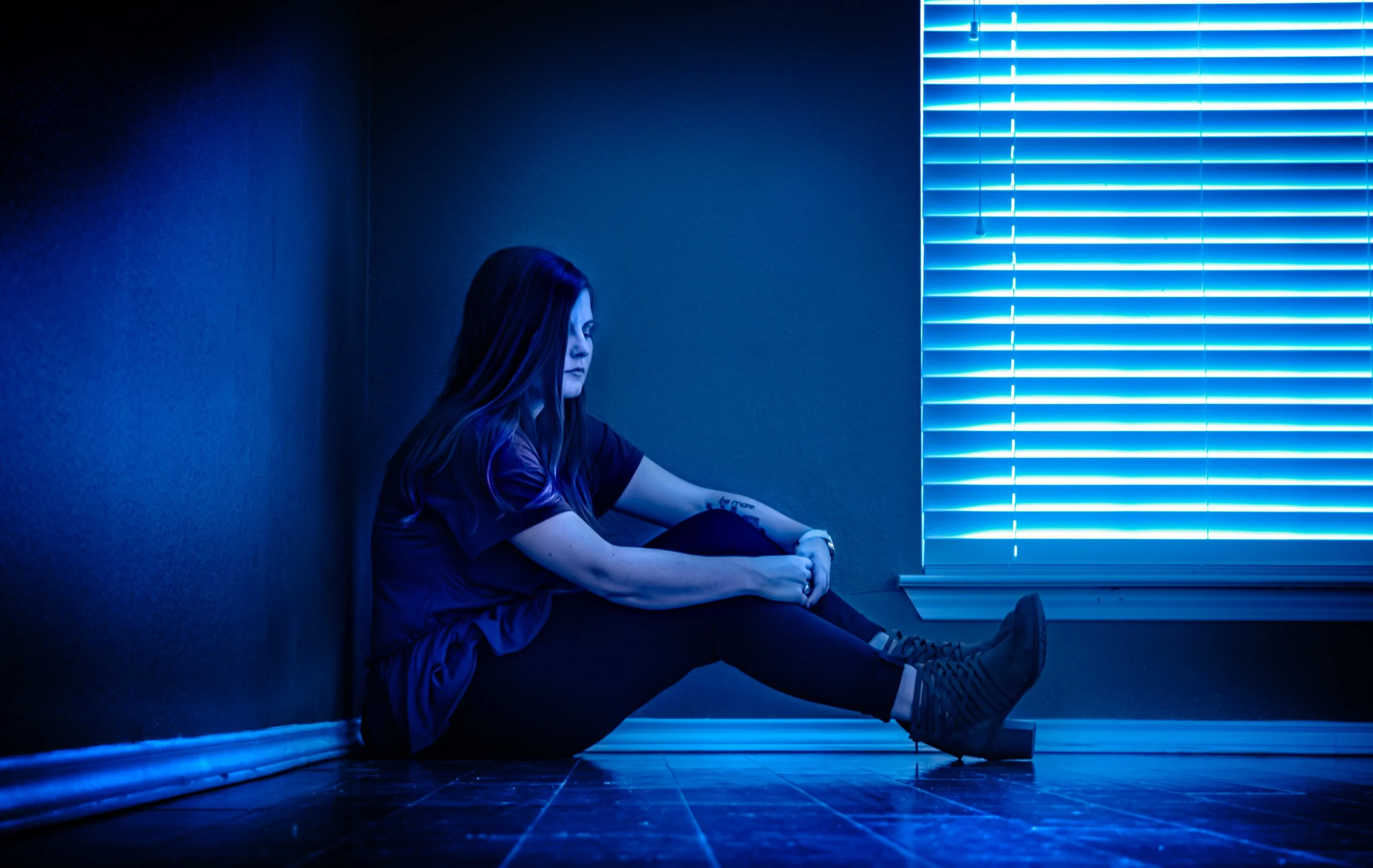 A young woman with long hair, dressed in a t-shirt and sneakers, sitting on the floor of a dimly lit room with blue neon lighting and closed blinds.