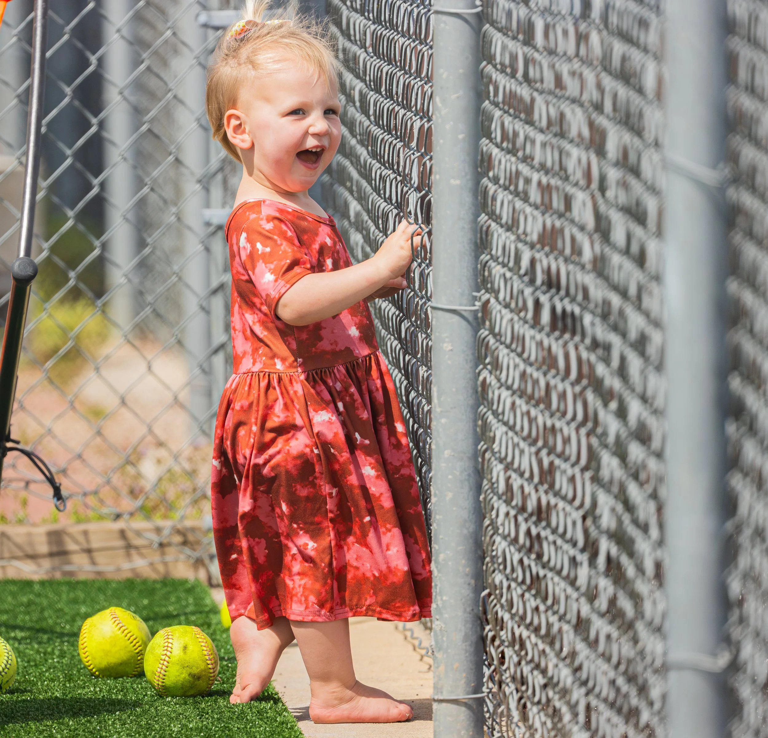A young girl with blonde hair wearing a red tie-dye dress standing barefoot near a chain-link fence, surrounded by yellow softballs on artificial grass.