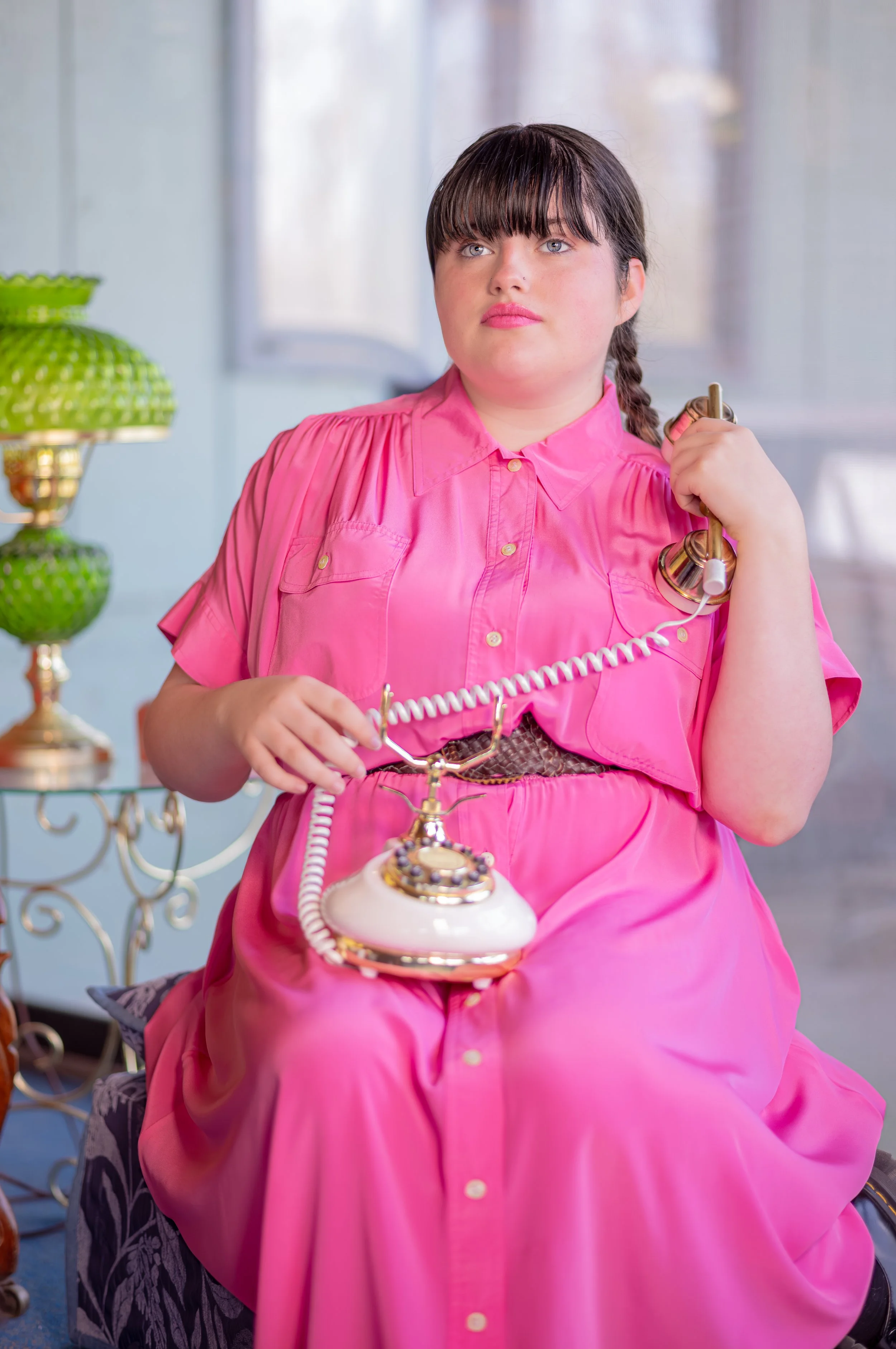 A woman with dark hair and bangs, wearing a bright pink button-up dress, sitting on a chair, holding a vintage rotary phone with a gold and cream color scheme, in a room with a lamp and a table with decorative items.