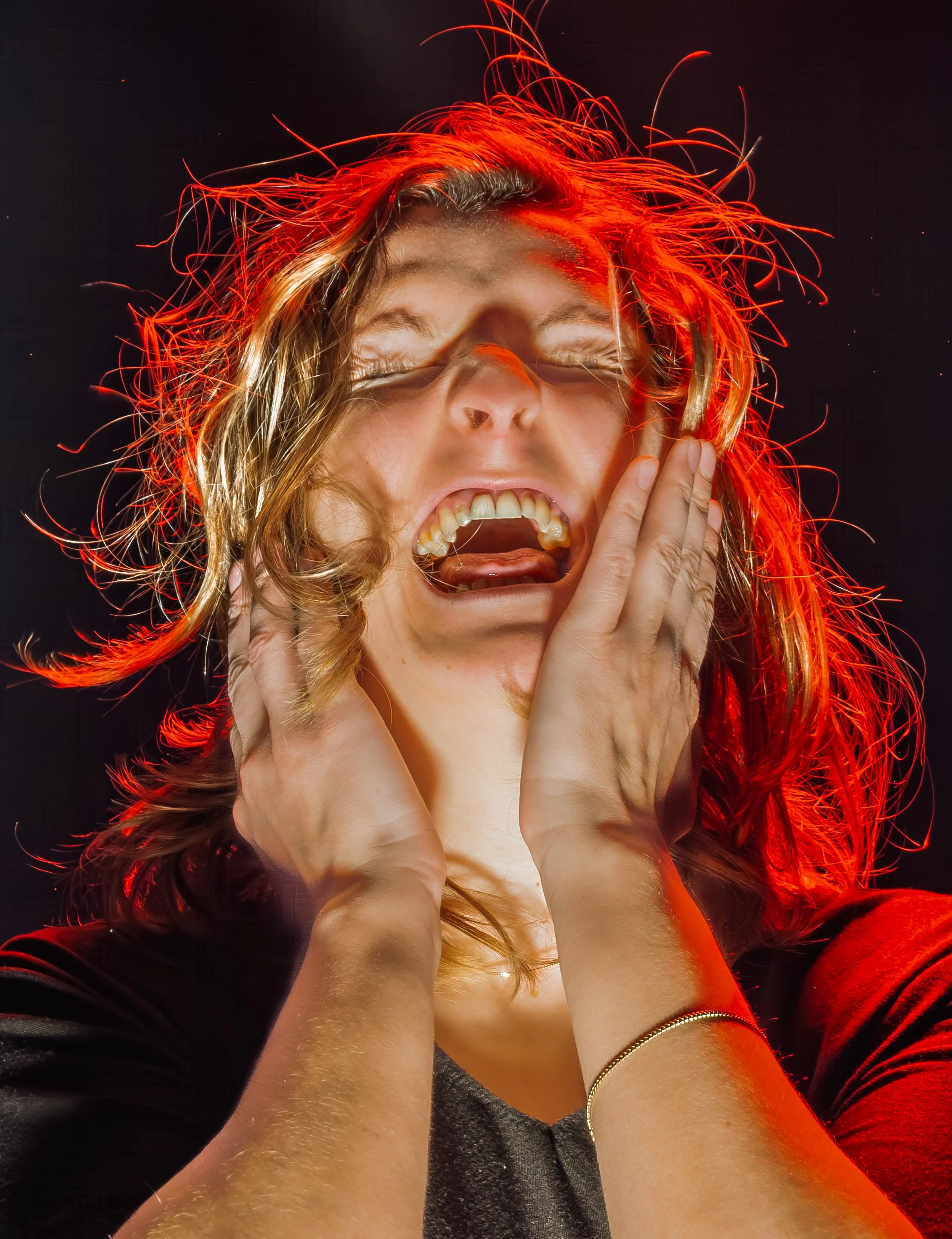 A woman with long wavy hair appears to be shouting or singing with her mouth open, hands on her face, and eyes closed, surrounded by red light streaks.