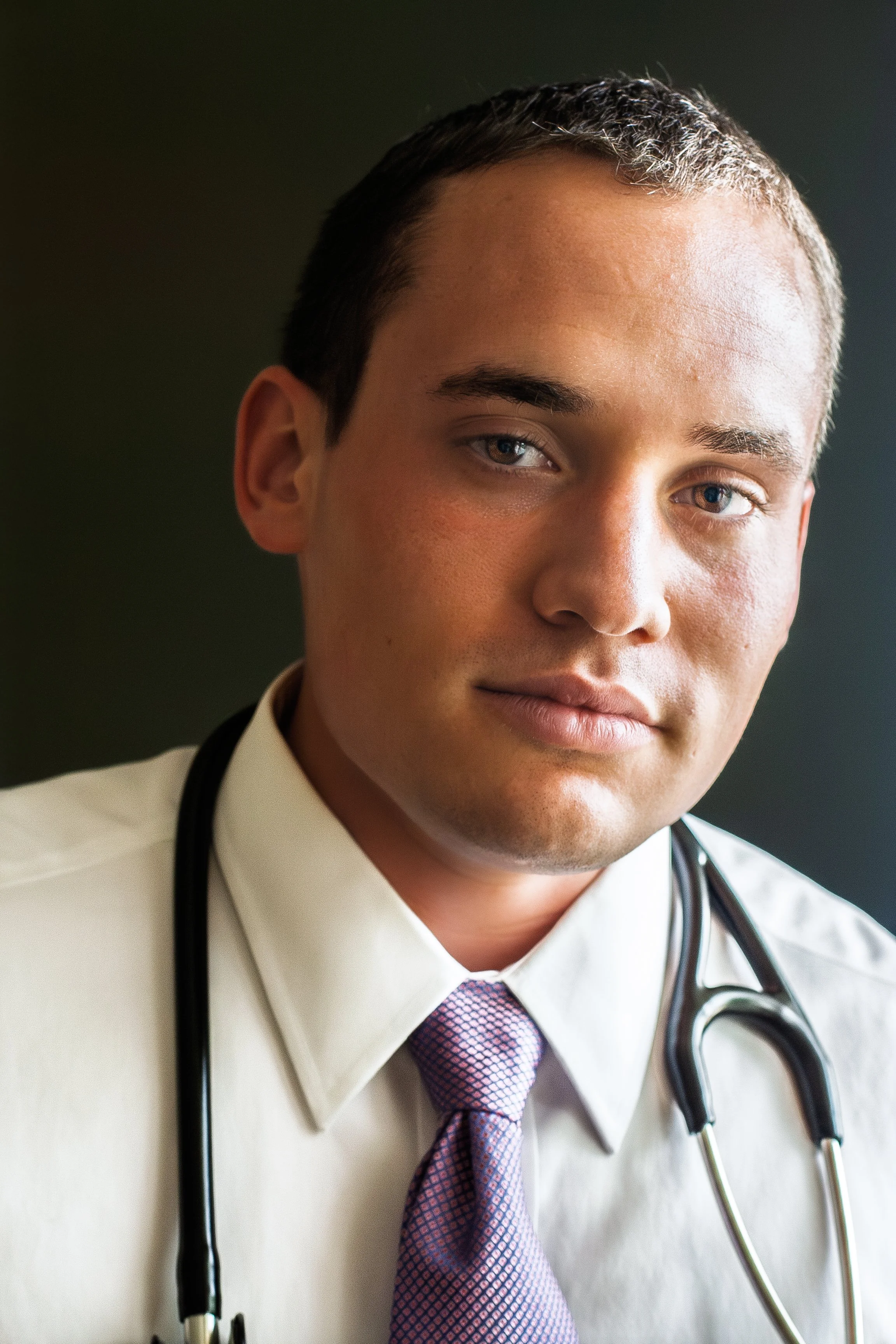 Close-up of a young male doctor with stethoscope around his neck, wearing a white shirt and a purple patterned tie, looking at the camera with a slight smile.