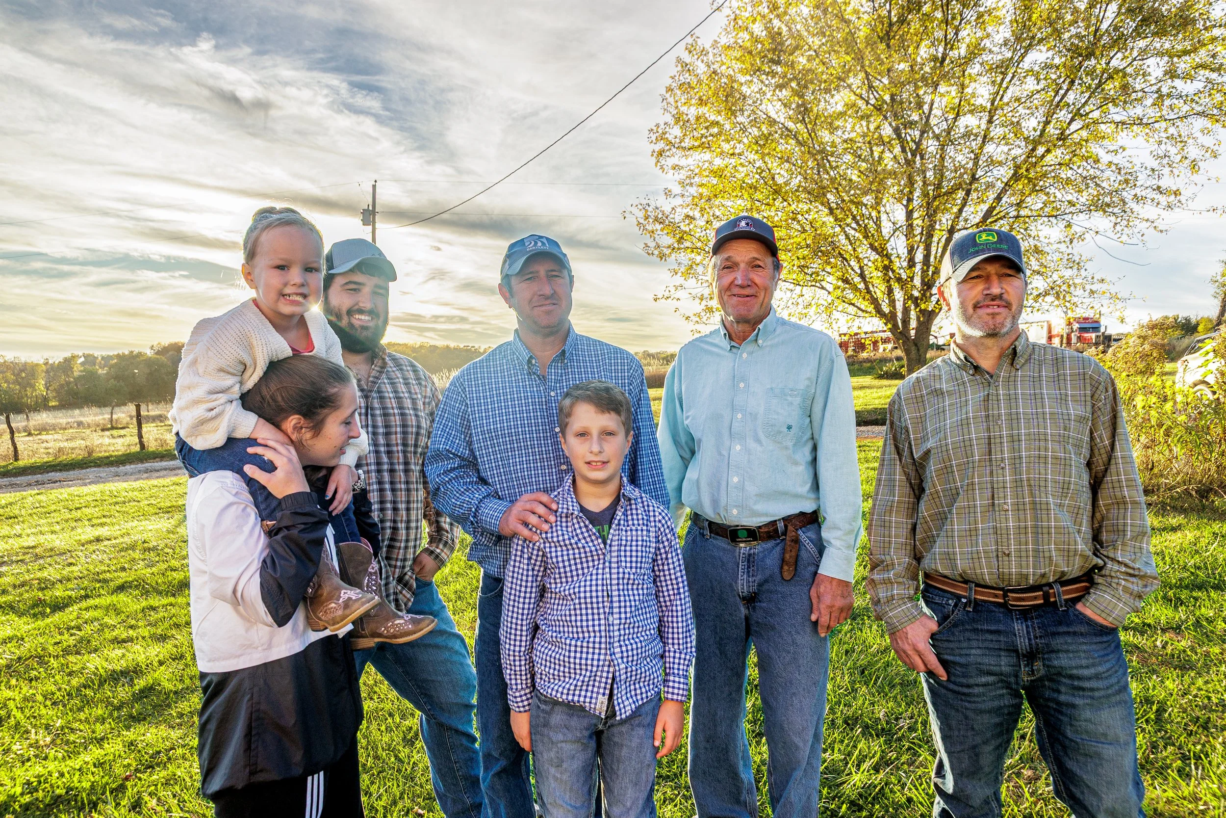 Group of seven people, including children and adults, gathered outdoors on a grassy area with trees and a cloudy sky in the background, during sunset or late afternoon.