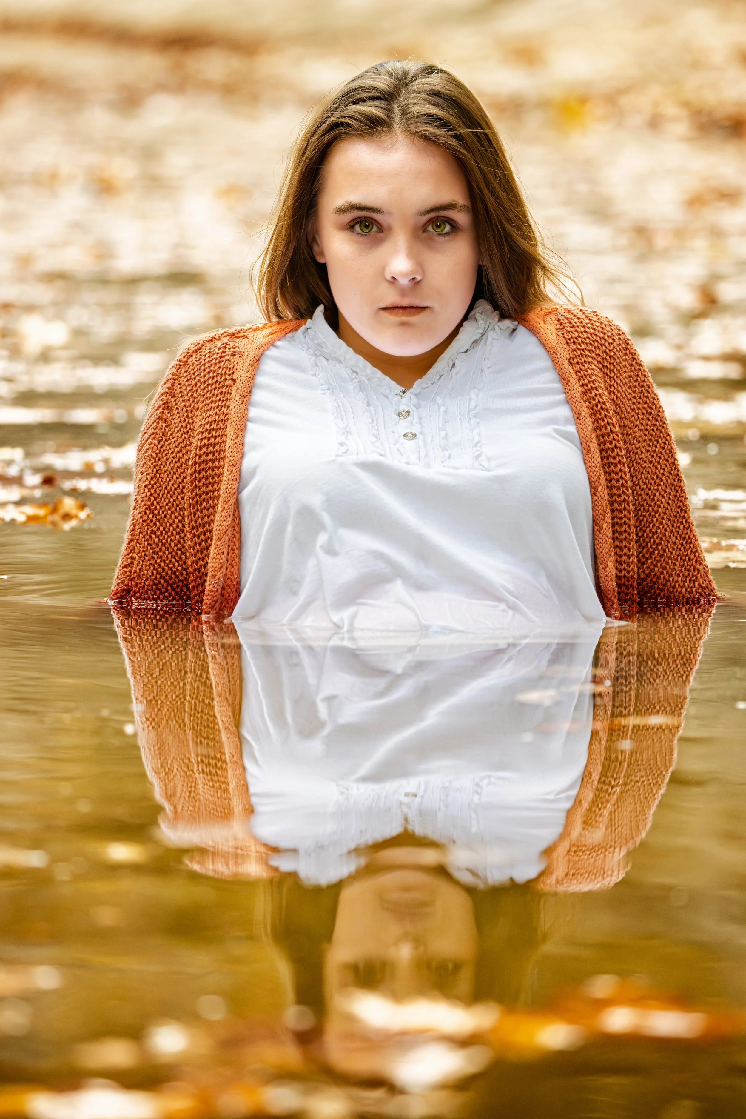 A young woman with brown hair and green eyes stands in a body of water, wearing a white shirt and an orange cardigan. She appears to be outdoors with autumn leaves floating on the water's surface.