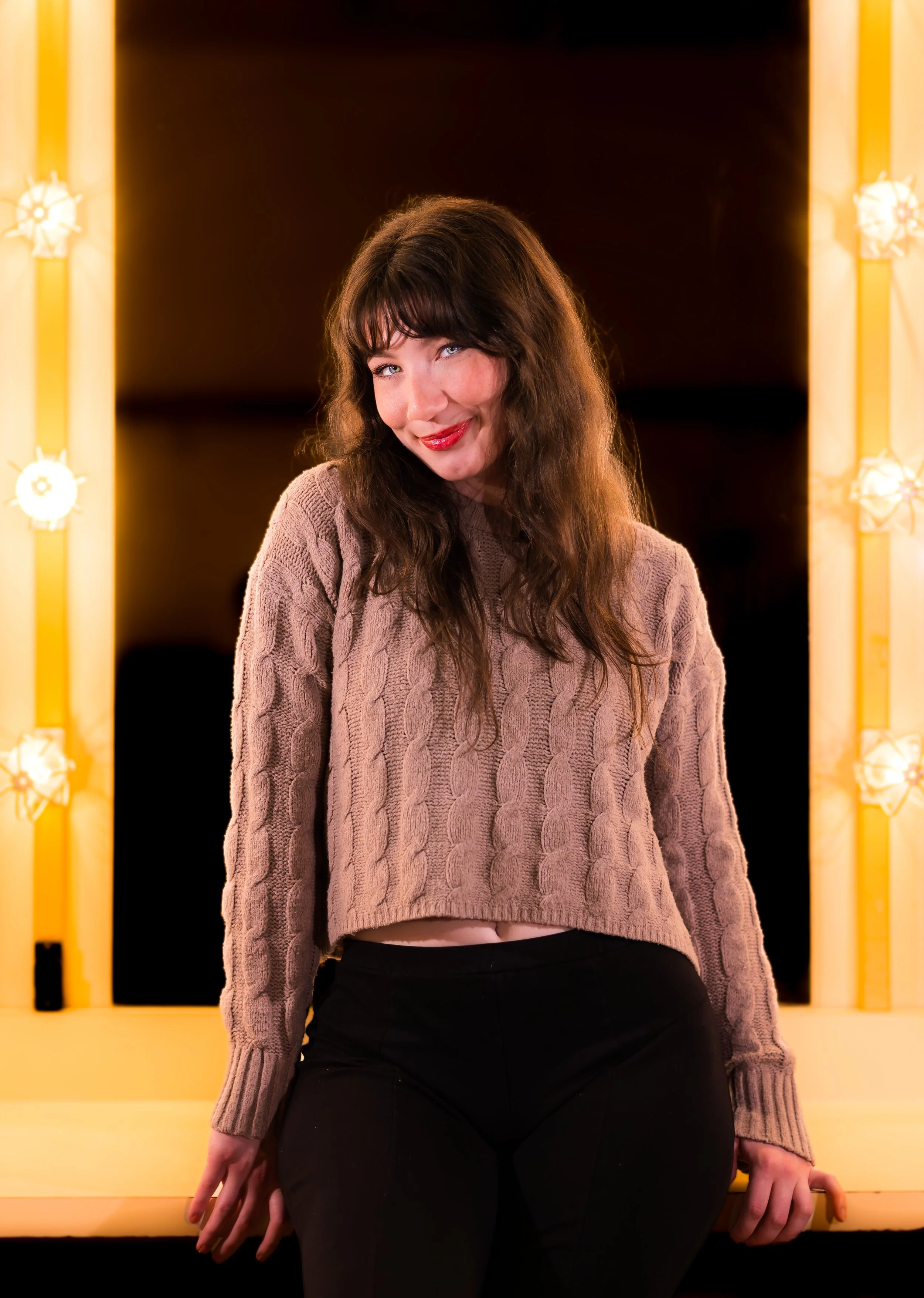 A woman with wavy brown hair and blue eyes, wearing a beige cable-knit sweater and black pants, sitting in front of a yellow-lit vanity mirror with a dark background.