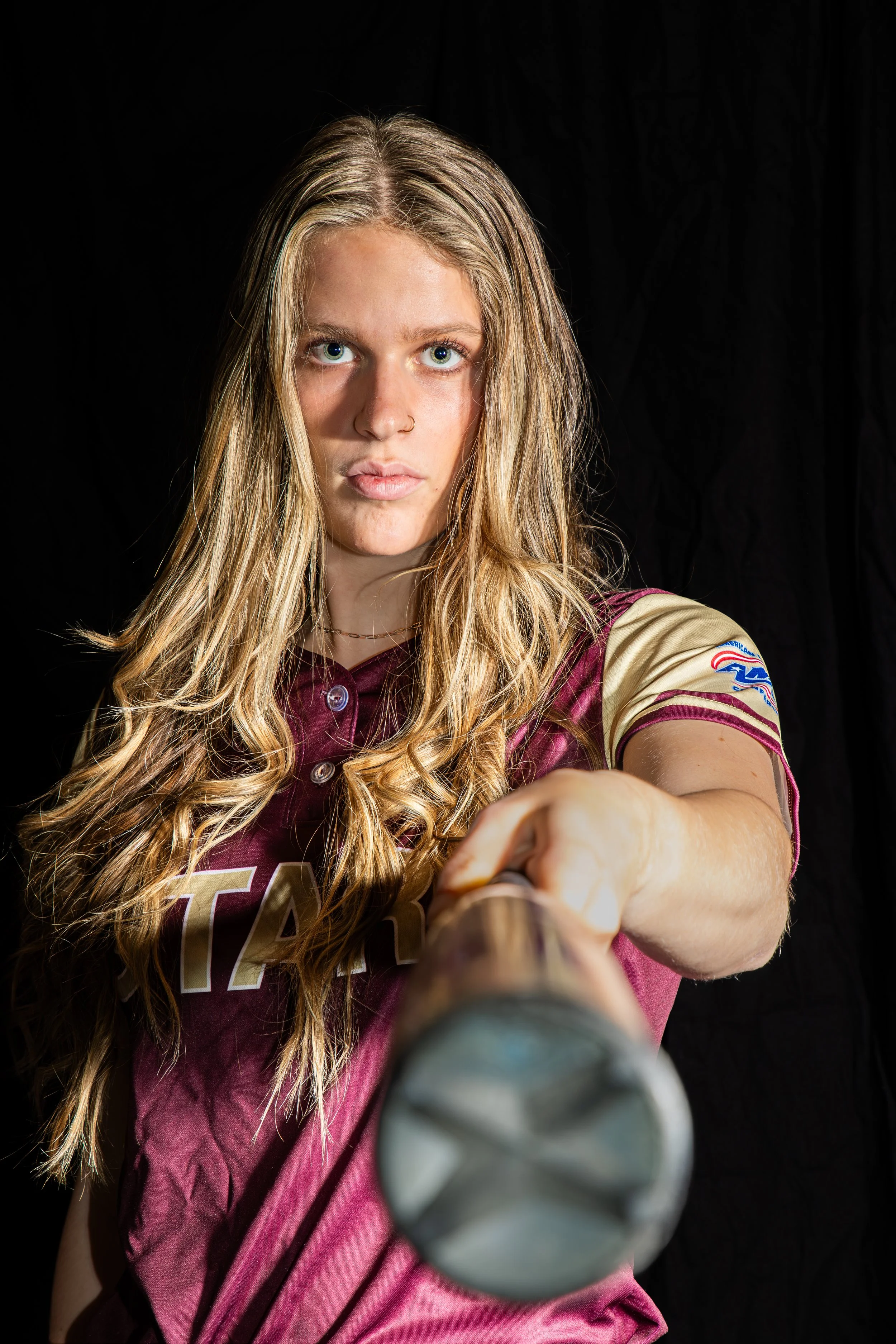 A young woman with long, wavy hair and blue eyes extending her hand forward holding a baseball bat, wearing a maroon sports jersey with gold and beige accents.