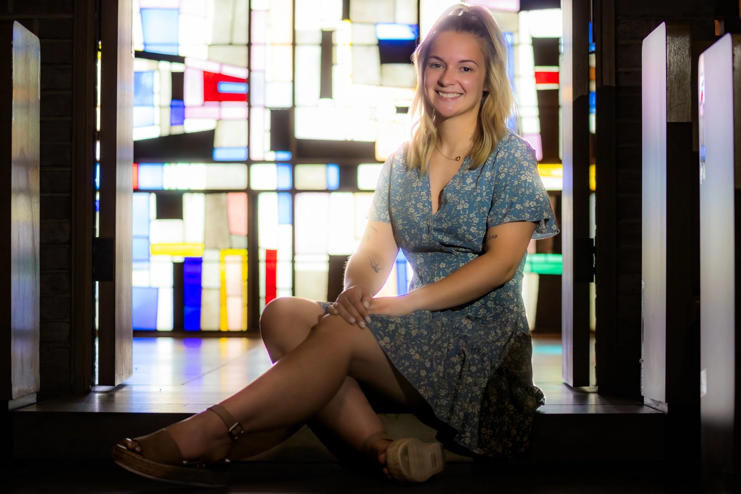 A young woman with blonde hair sitting on the floor in front of a colorful stained glass window, smiling at the camera. She is wearing a blue floral dress, tan sandals, and has a tattoo on her arm.