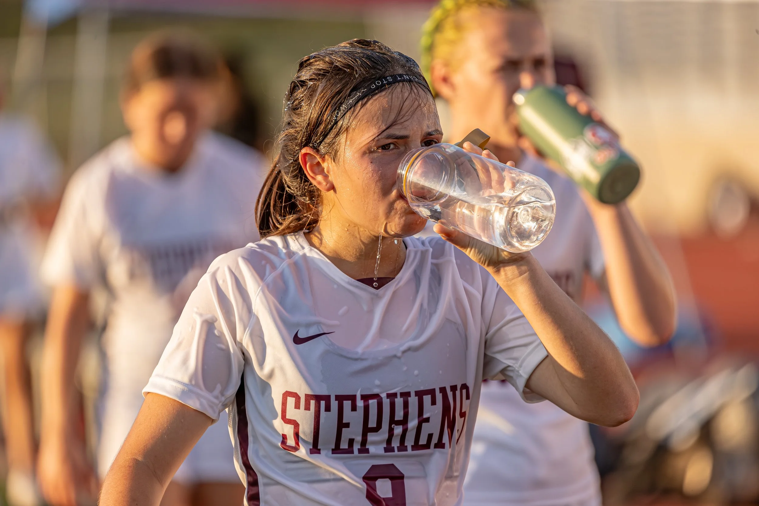 A female soccer player in a white and red uniform drinking water from a clear water bottle after a game or practice, with other players in the background.