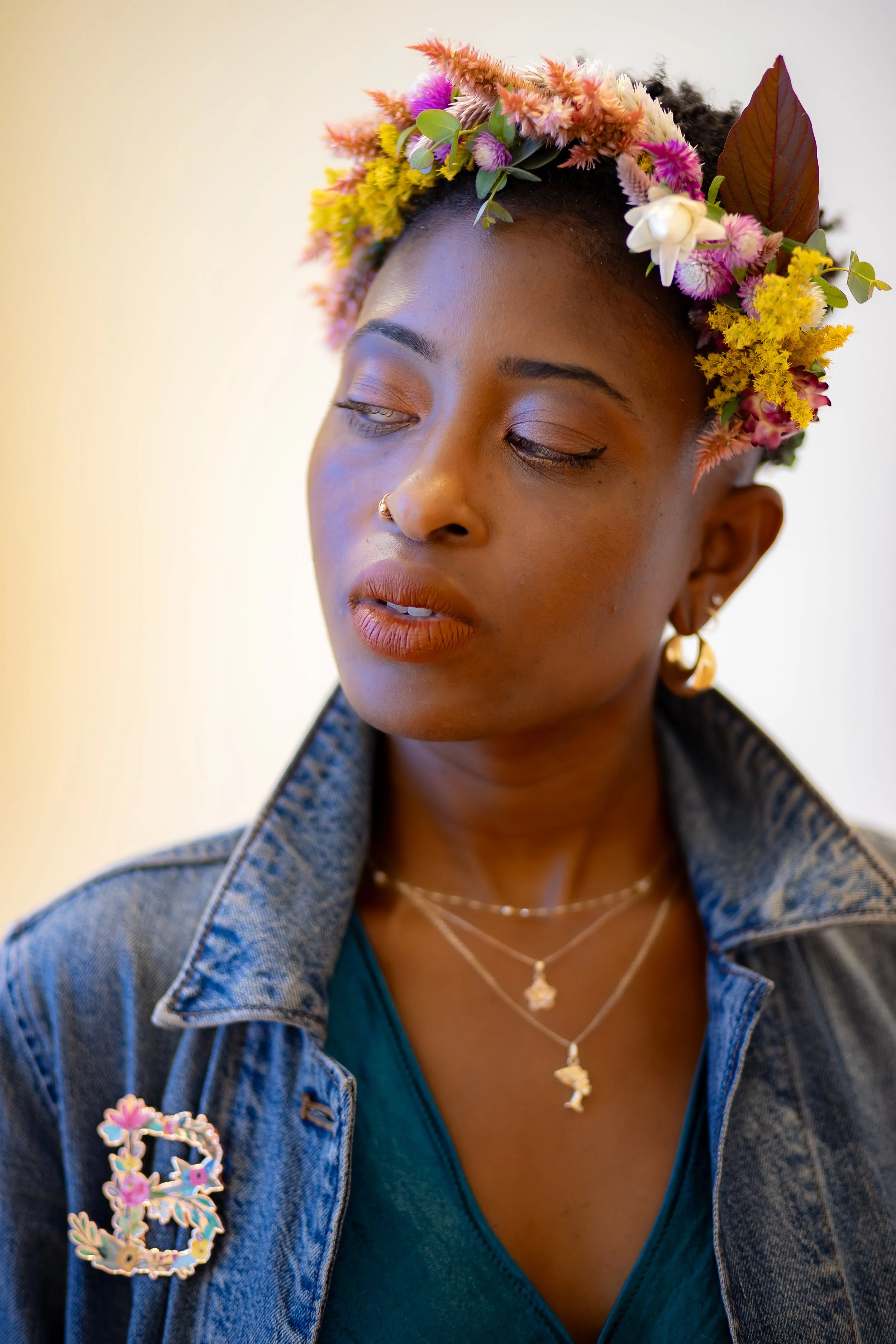 A woman with closed eyes wearing a floral crown, earrings, necklaces, a denim jacket, and blue top.