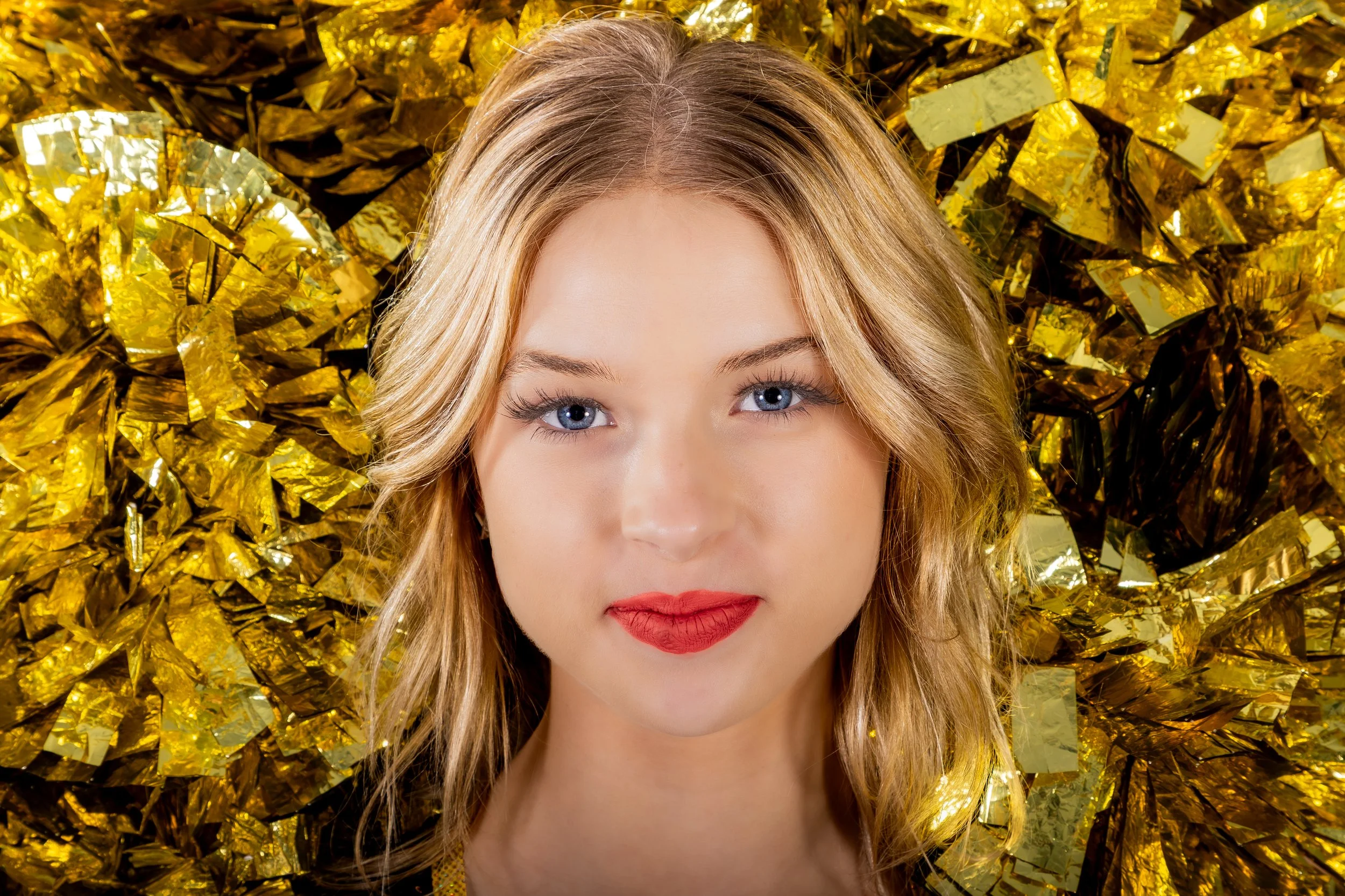 A woman with blonde hair and blue eyes lying on a bed of gold foil or metallic paper. She has red lipstick and is looking directly at the camera.