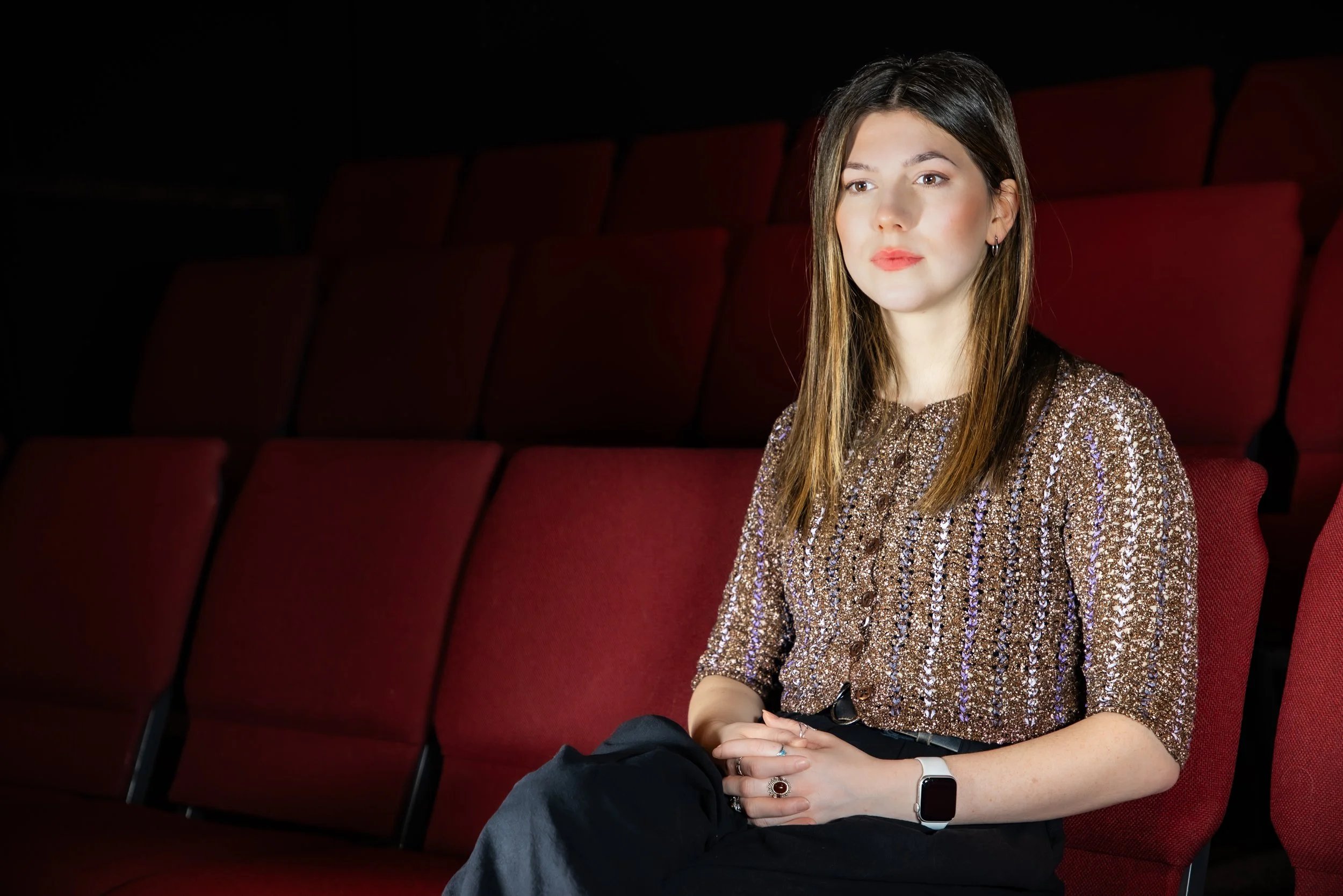 A woman with long brown hair sitting in a theater with red seats, wearing a shiny, patterned top and black pants.