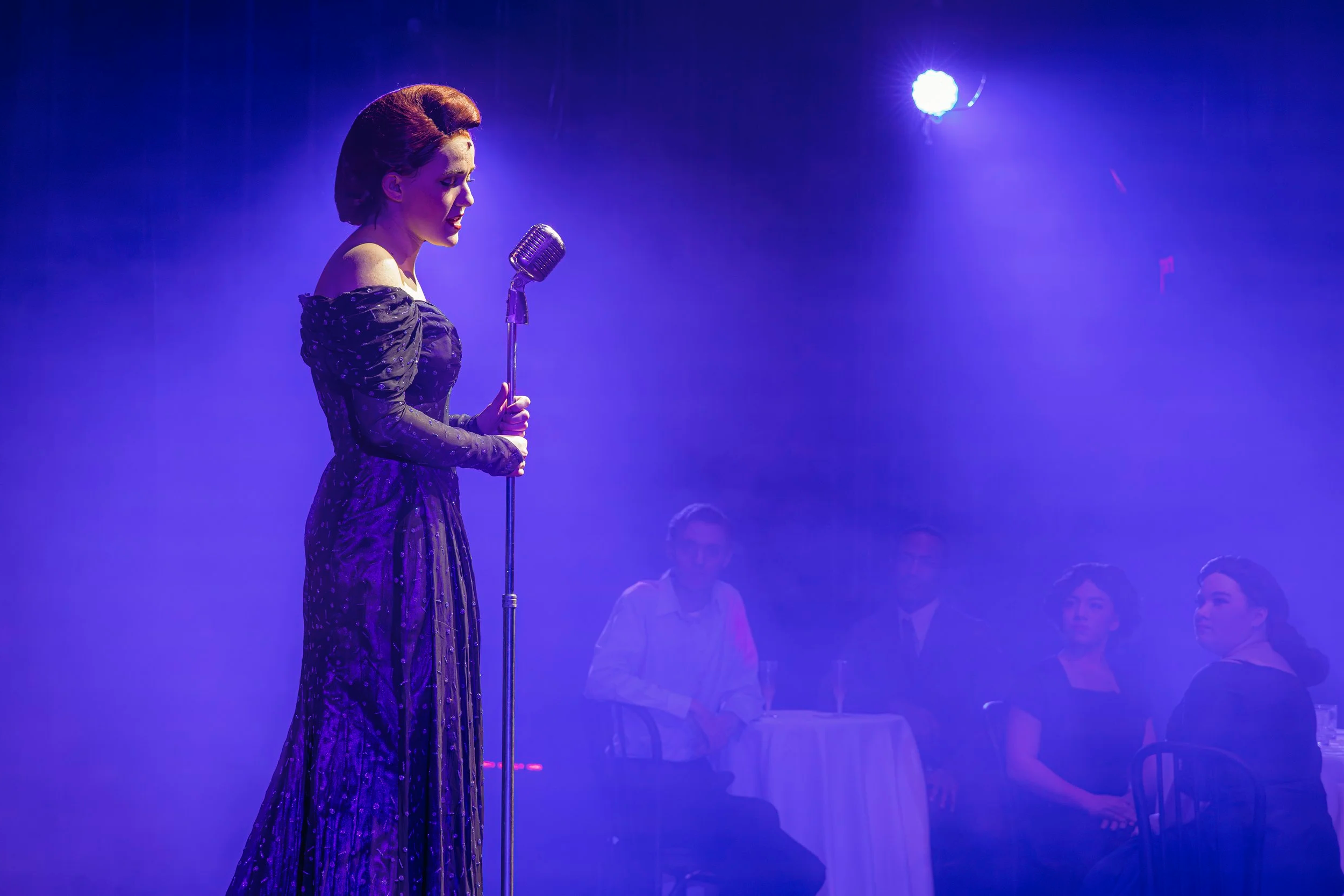 A woman in a purple vintage dress singing on stage with a microphone, illuminated by purple stage lighting, with four people sitting at a table watching her.