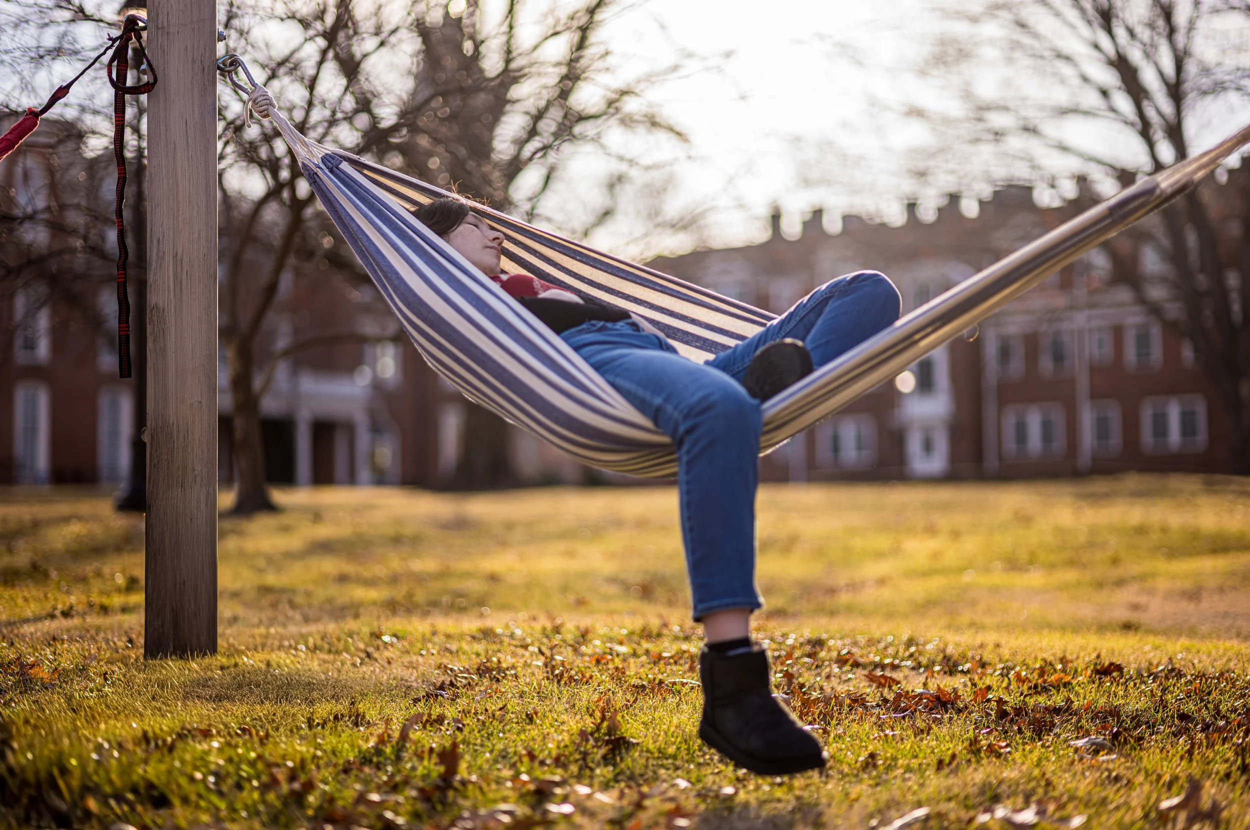 A person relaxing in a hammock outdoors on a sunny day, with trees and building in the background.