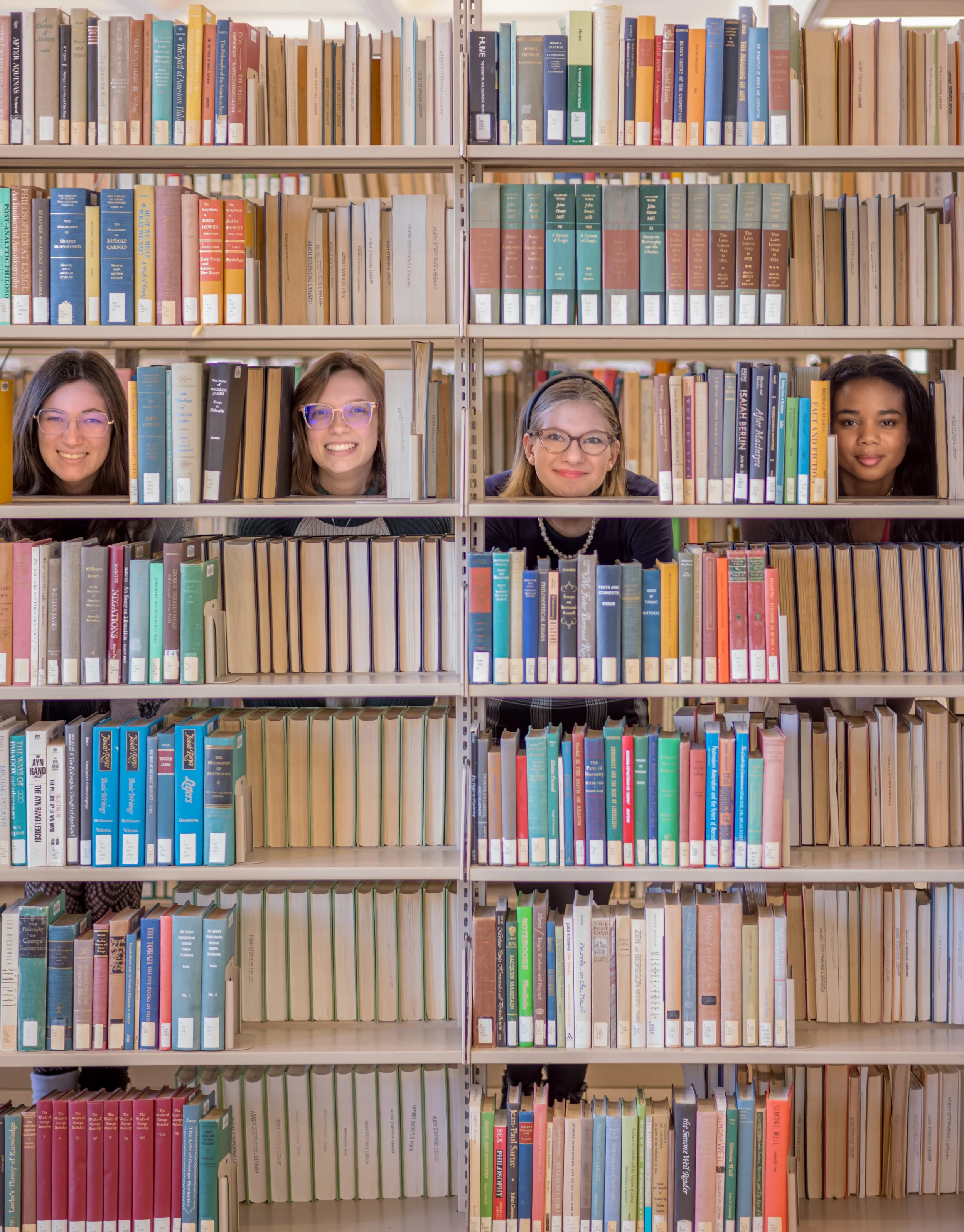 Four women peeking through library bookshelves, smiling, with numerous books in the background.