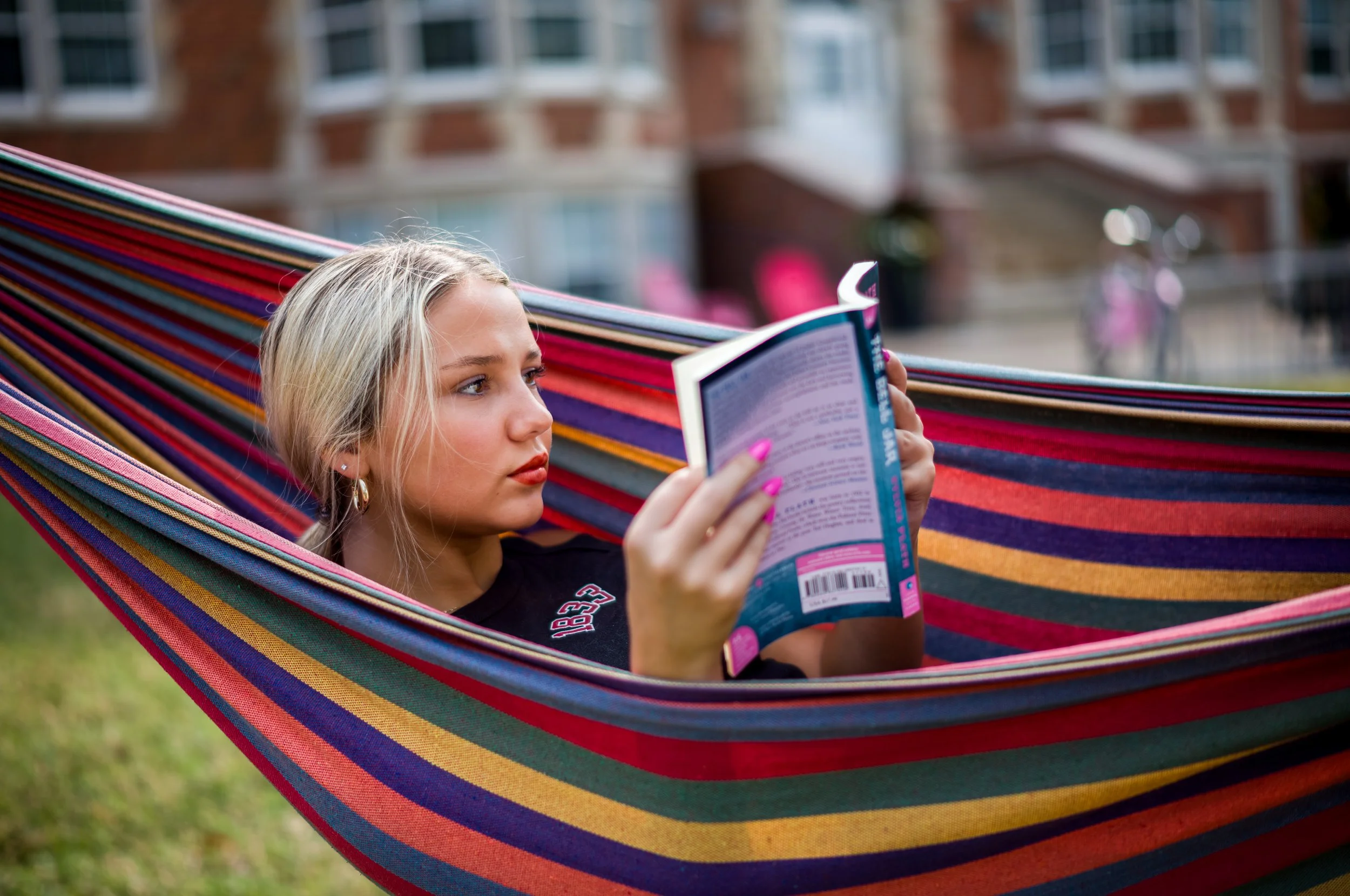 A young woman with blonde hair lying in a colorful striped hammock, reading a book outdoors with blurred buildings in the background.