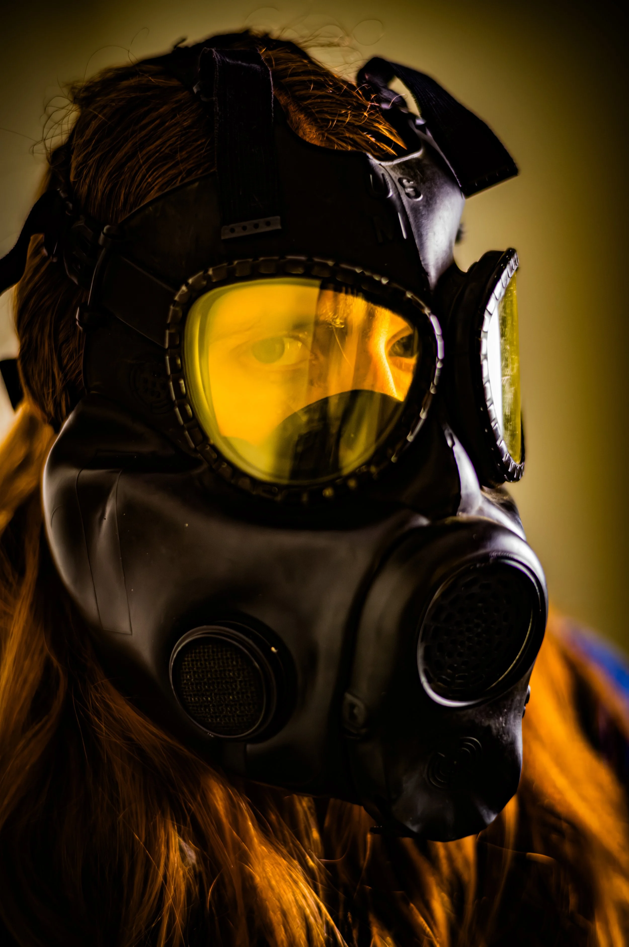 A person with long hair wearing a black gas mask and yellow-tinted goggles.