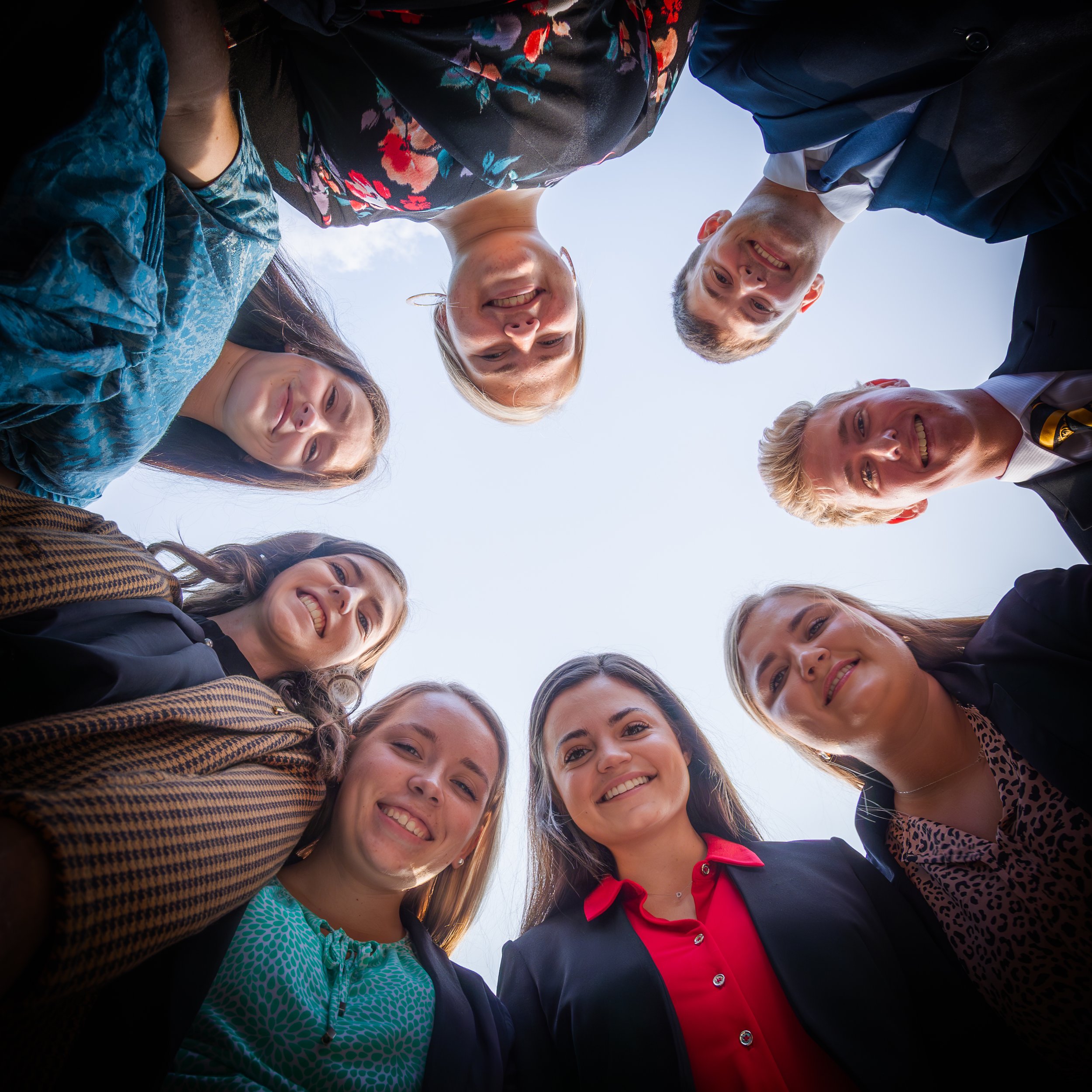 A group of smiling people, both men and women, standing in a circle outdoors looking down at the camera with the sky visible above them.