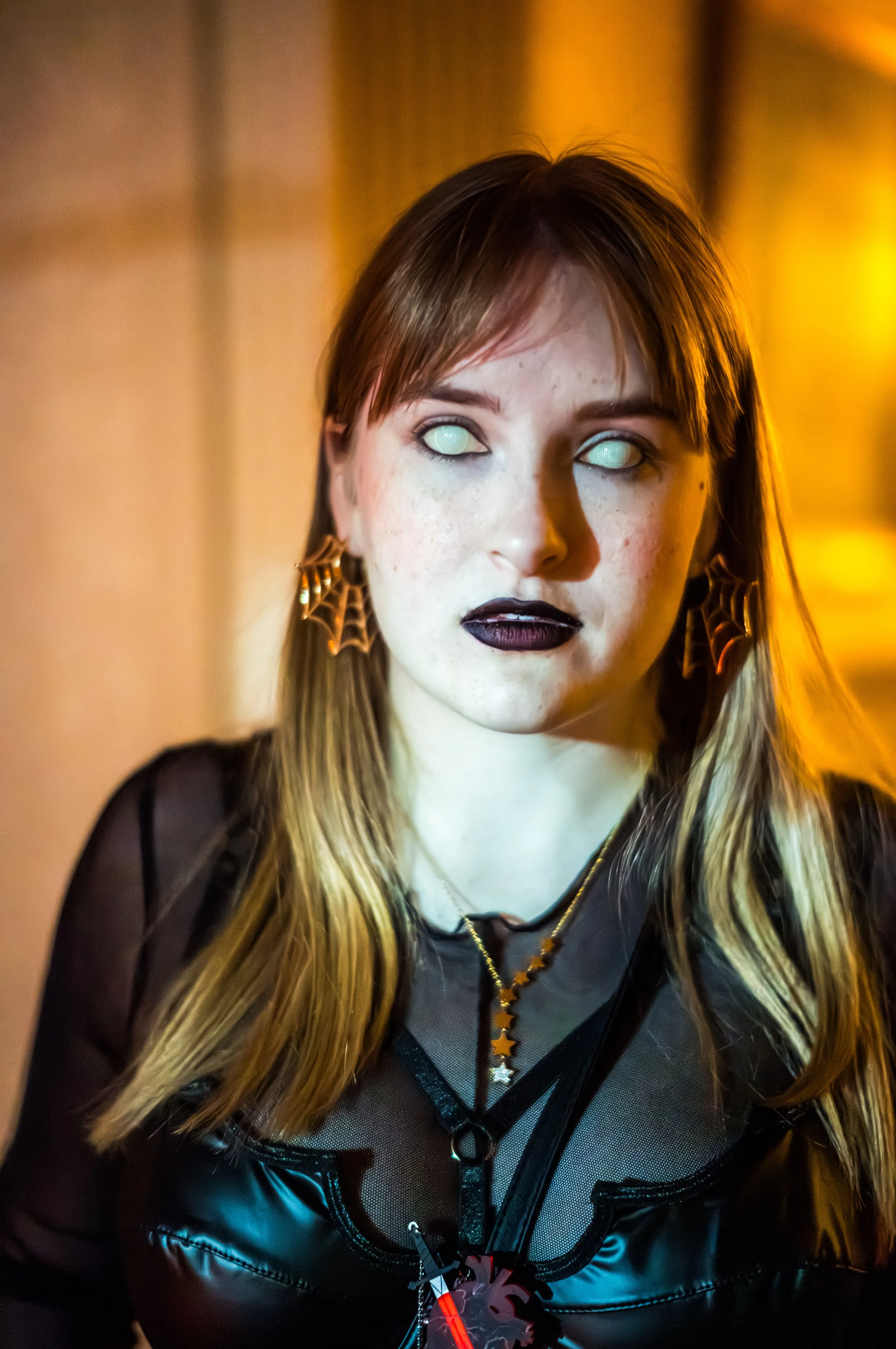 A person with long blonde hair, wearing black goth makeup with black lipstick, and ghostly white eyes, dressed in a black mesh top and leather jacket with dragon-shaped earrings and star-shaped necklace, standing against a warm yellow illuminated bac