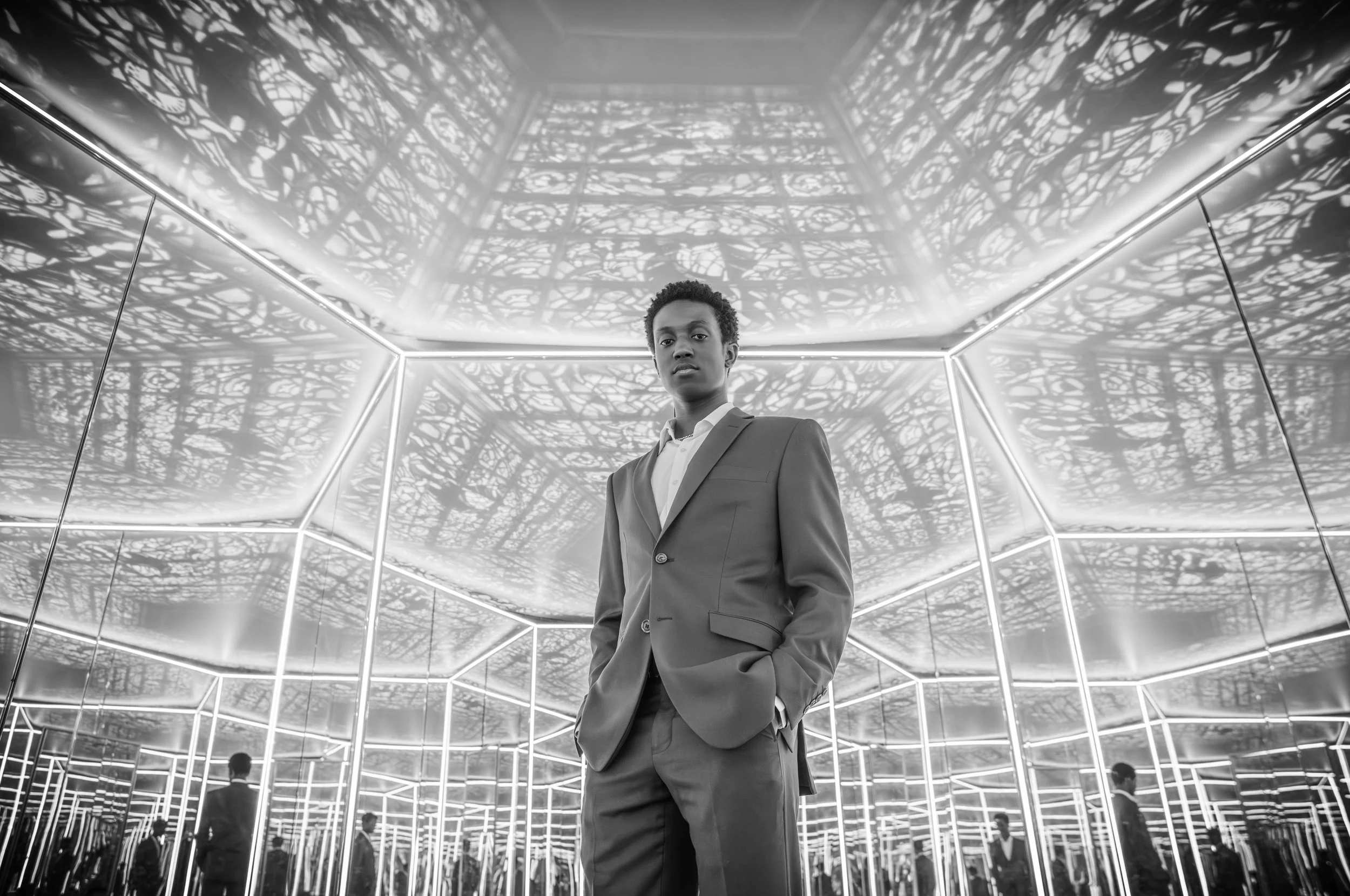 A woman in a business suit standing in a futuristic, mirrored room with illuminated edges and intricate ceiling patterns, with reflections of herself and others visible.