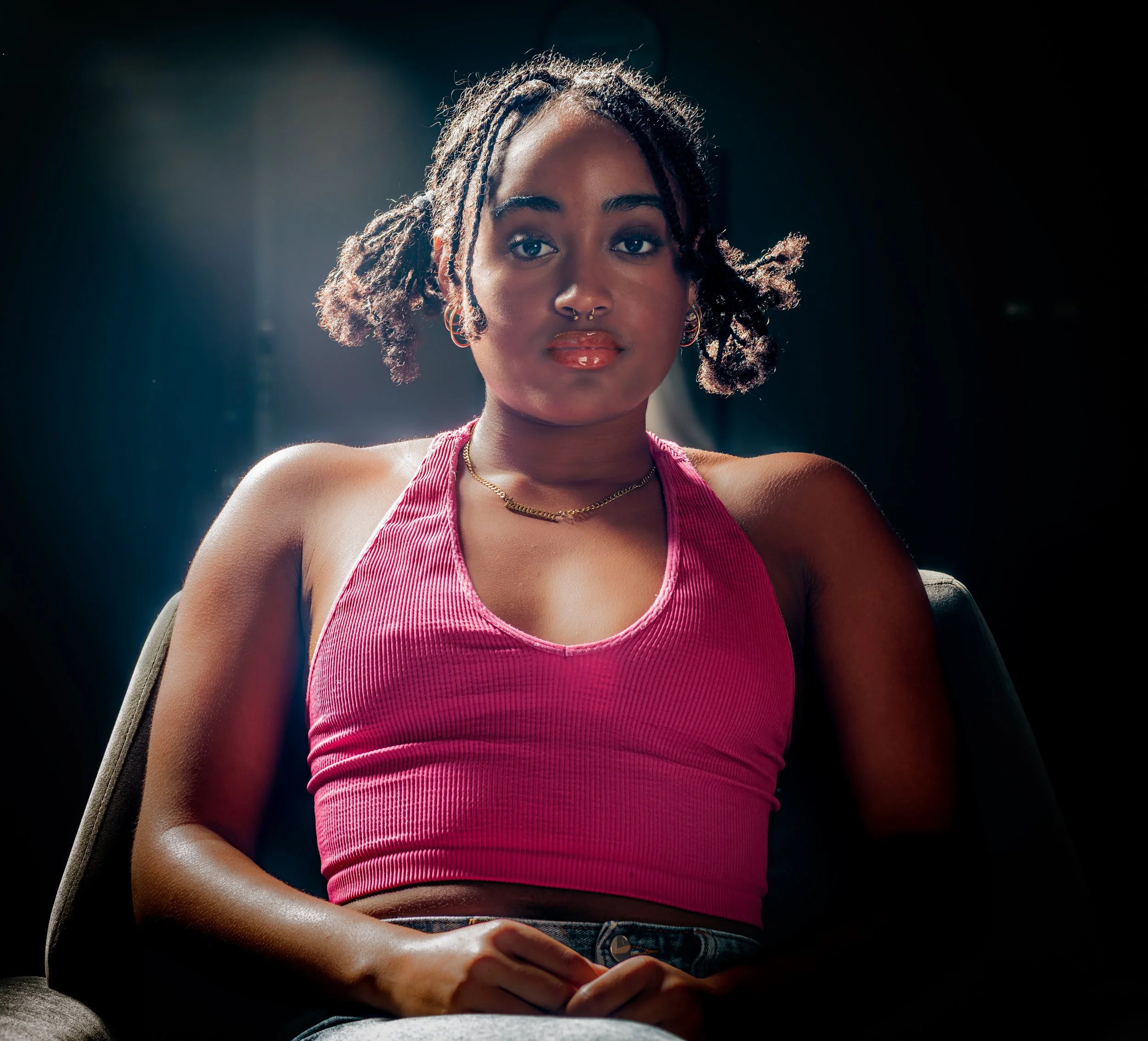 A young woman with braided hair and hoop earrings sitting with her hands on her lap, wearing a pink top, against a dark background.