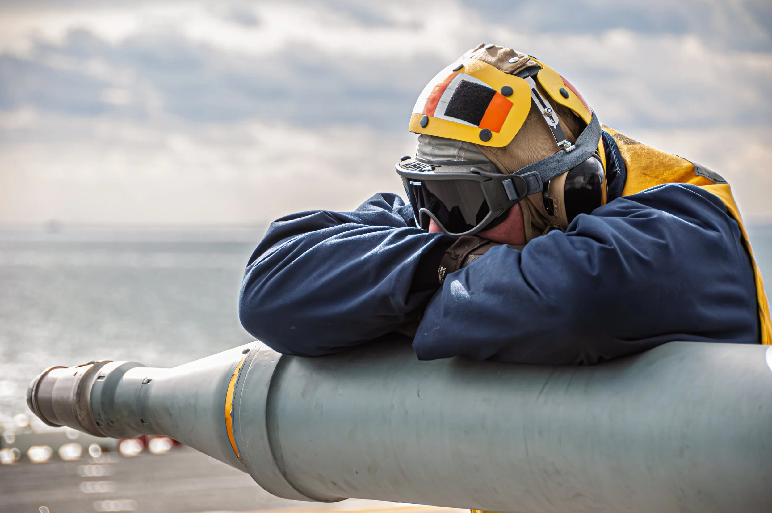 A person wearing a beige helmet, black goggles, a yellow and beige jacket, and blue gloves leaning on a large cylindrical object, with a hazy outdoor background.